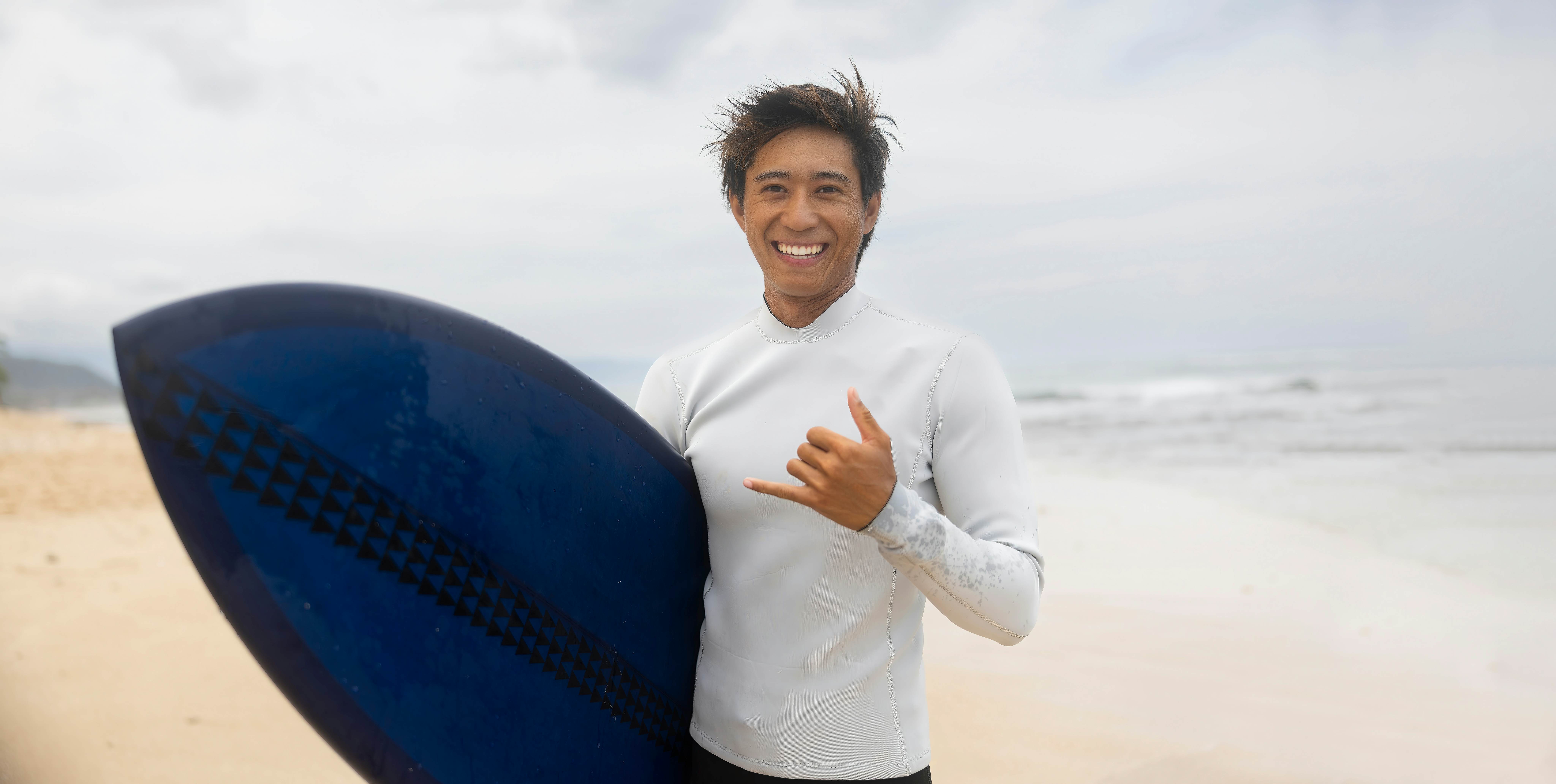 A surfer carrying a blue board giving the shaka hand gesture