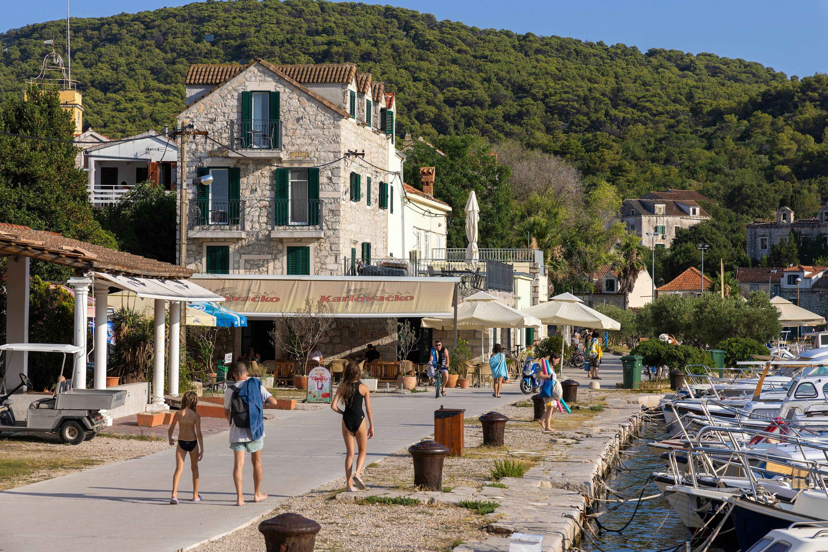 People walk on street by a port in a small town. Boats are moored along the quay to the right of the street.