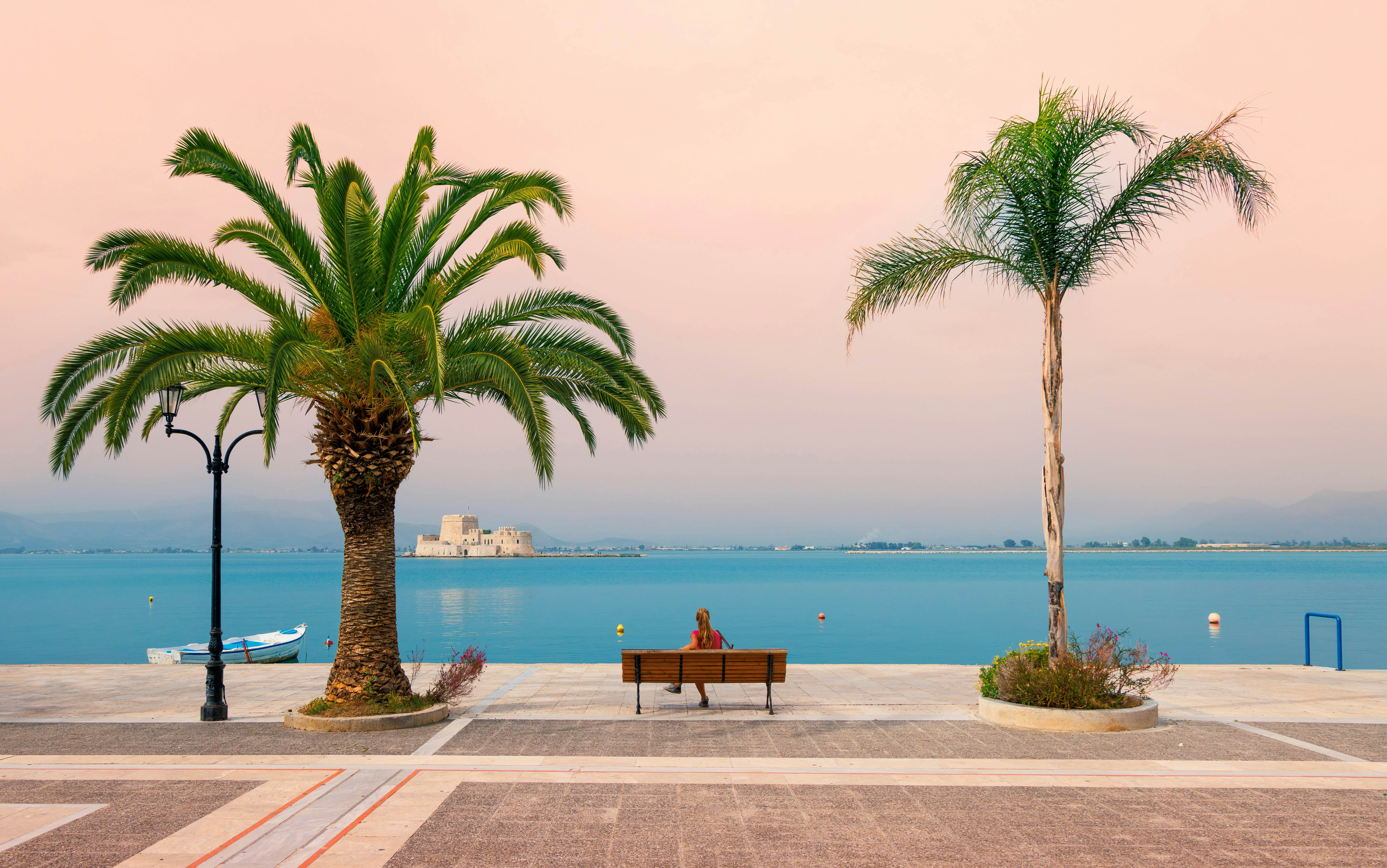 A person sits on a bench on a waterfront promenade, framed by two palm trees. An island fortress can be seen in the distance over the still blue water.