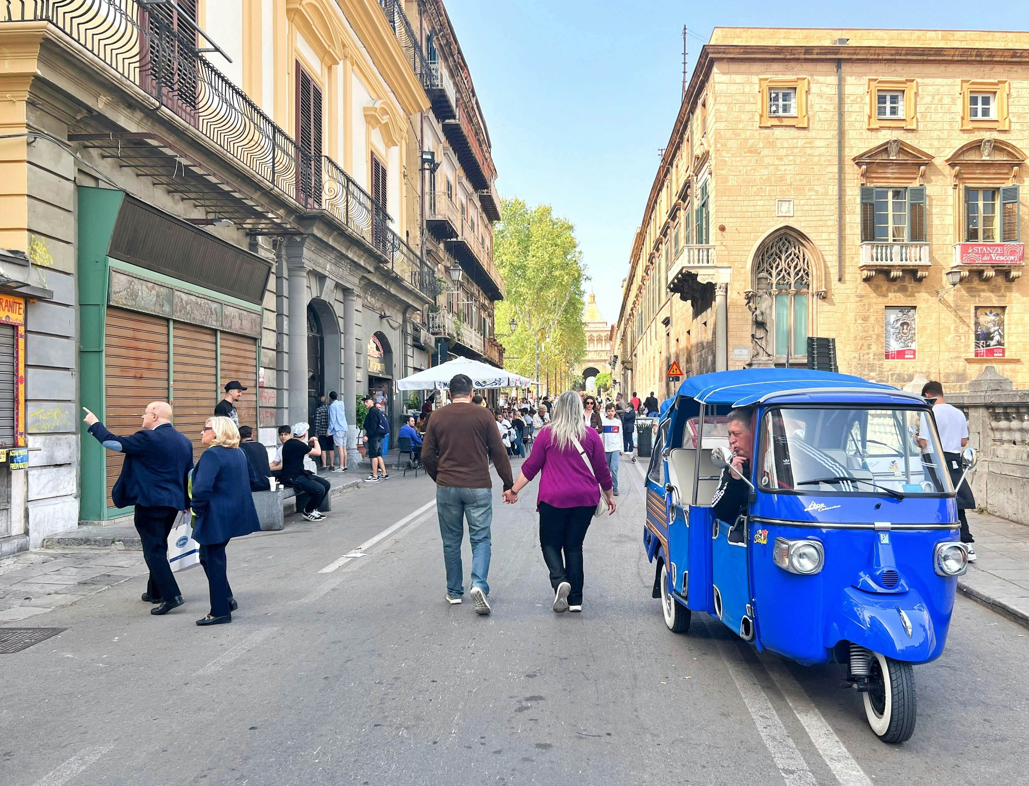 People walk down a street in a historic city. The street is closed to traffic – except for a blue, three-wheeled taxi in the foreground of the image.