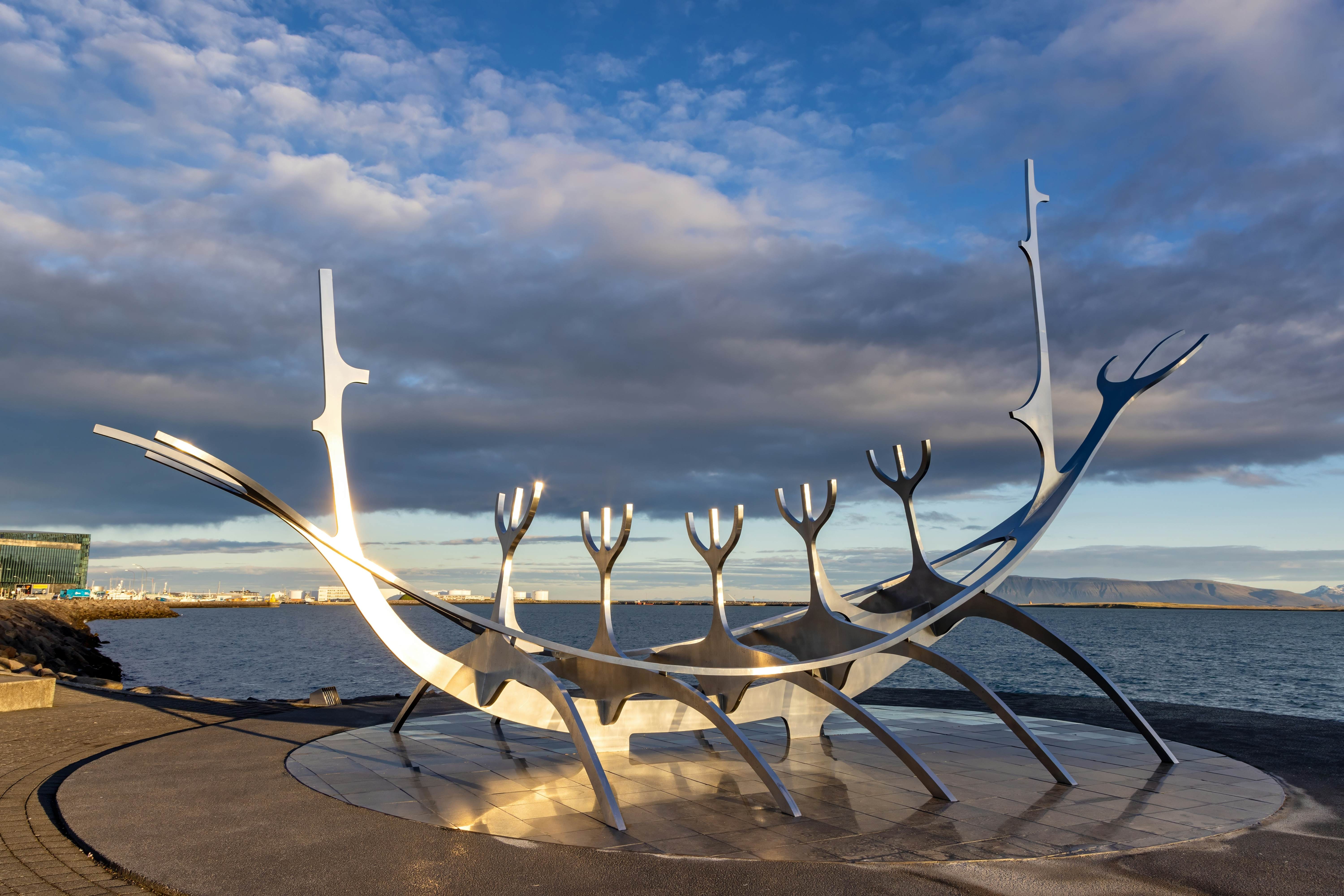 A public sculpture on a seafront plaza in a city depicts an abstracted Viking ship. The metal of the sculpture is illuminated by sun beams.