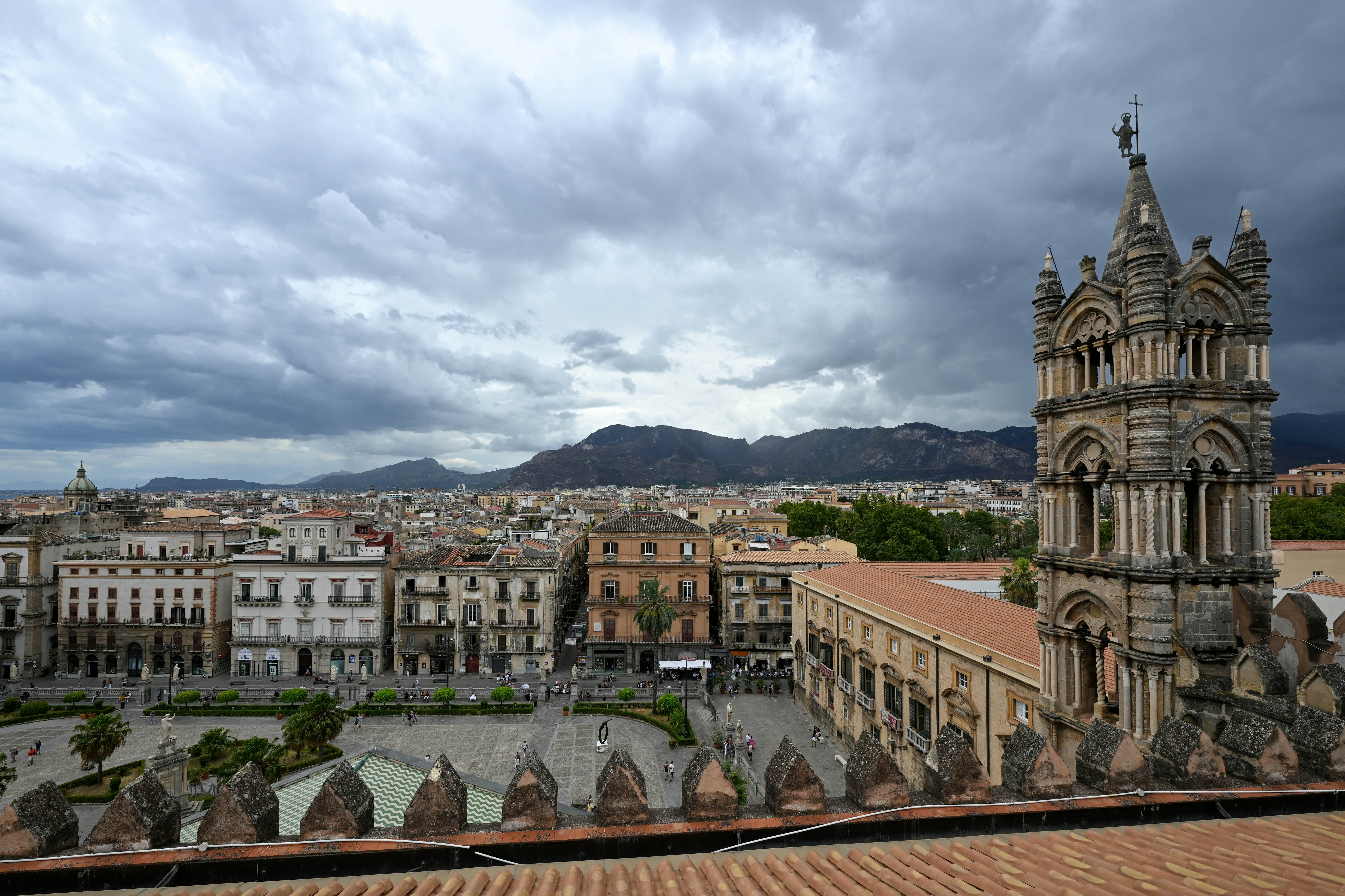 A view from a roof of a square in a European city. To the right, a medieval cathedral tower rises above red-tiled roofs.