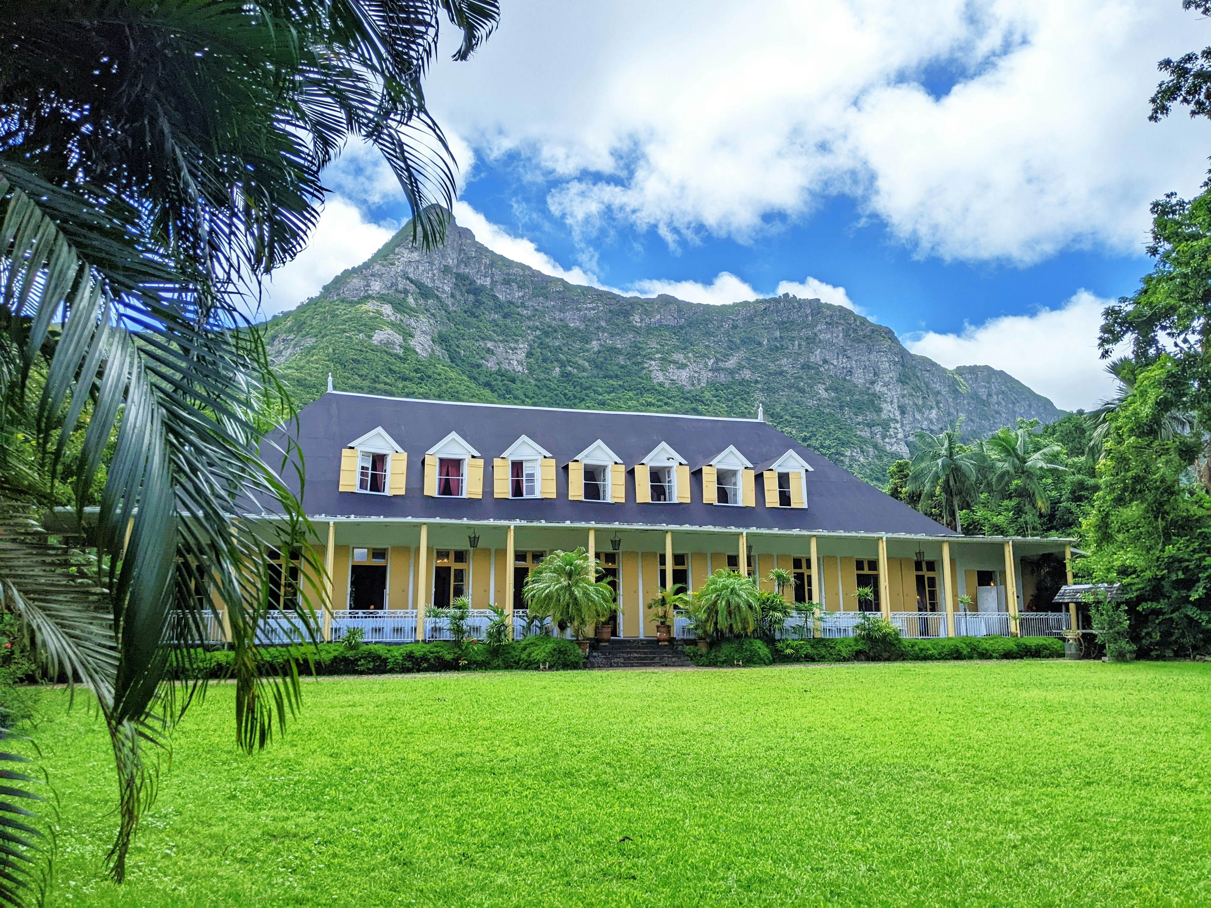 A colonial-style house with a long covered porch and dormers in front of a tidy lawn. A large hill looms behind the house.