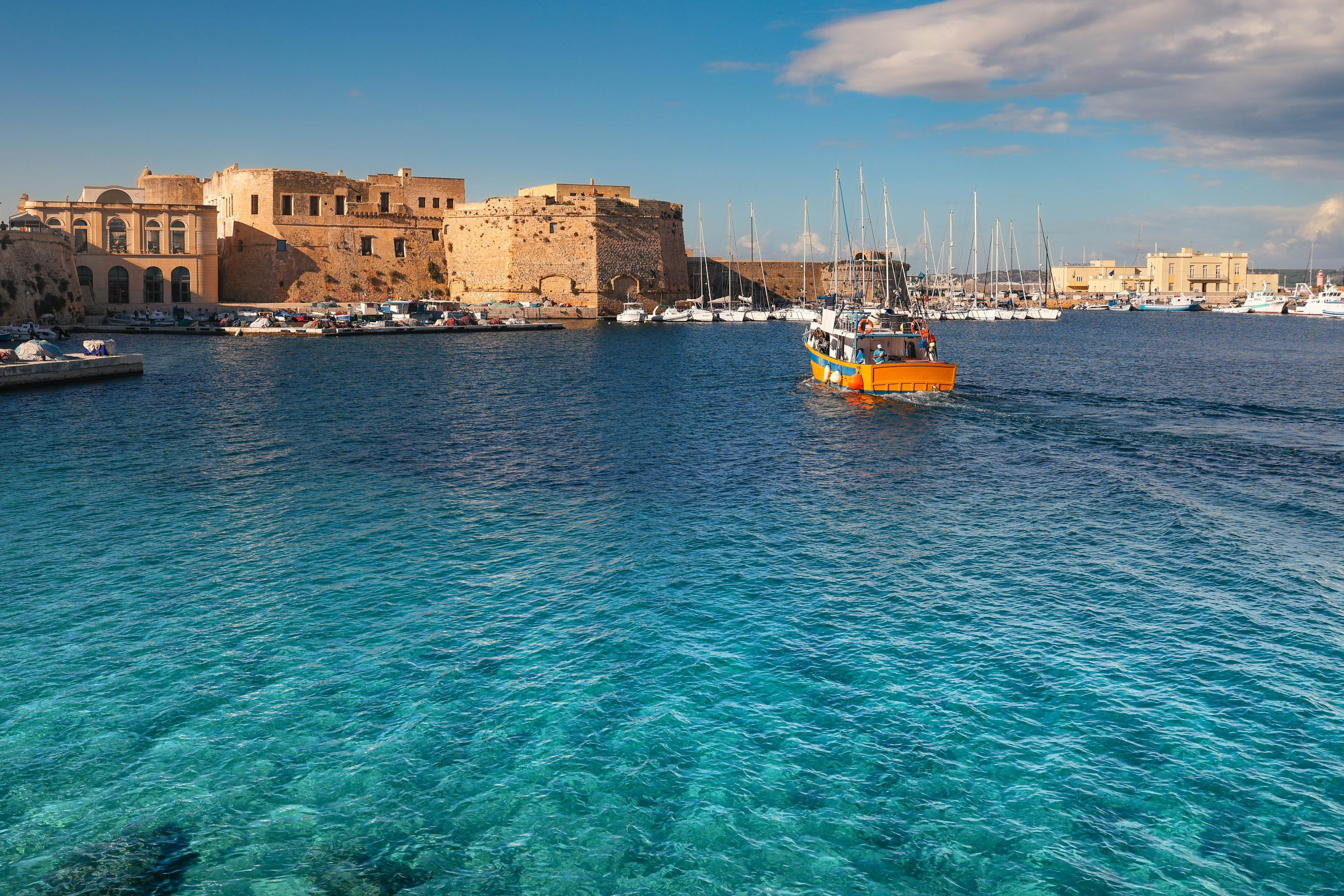 An ancient coastal city seen from sea, with a fortified castle, yacht marina and fishing boat on sunny day.