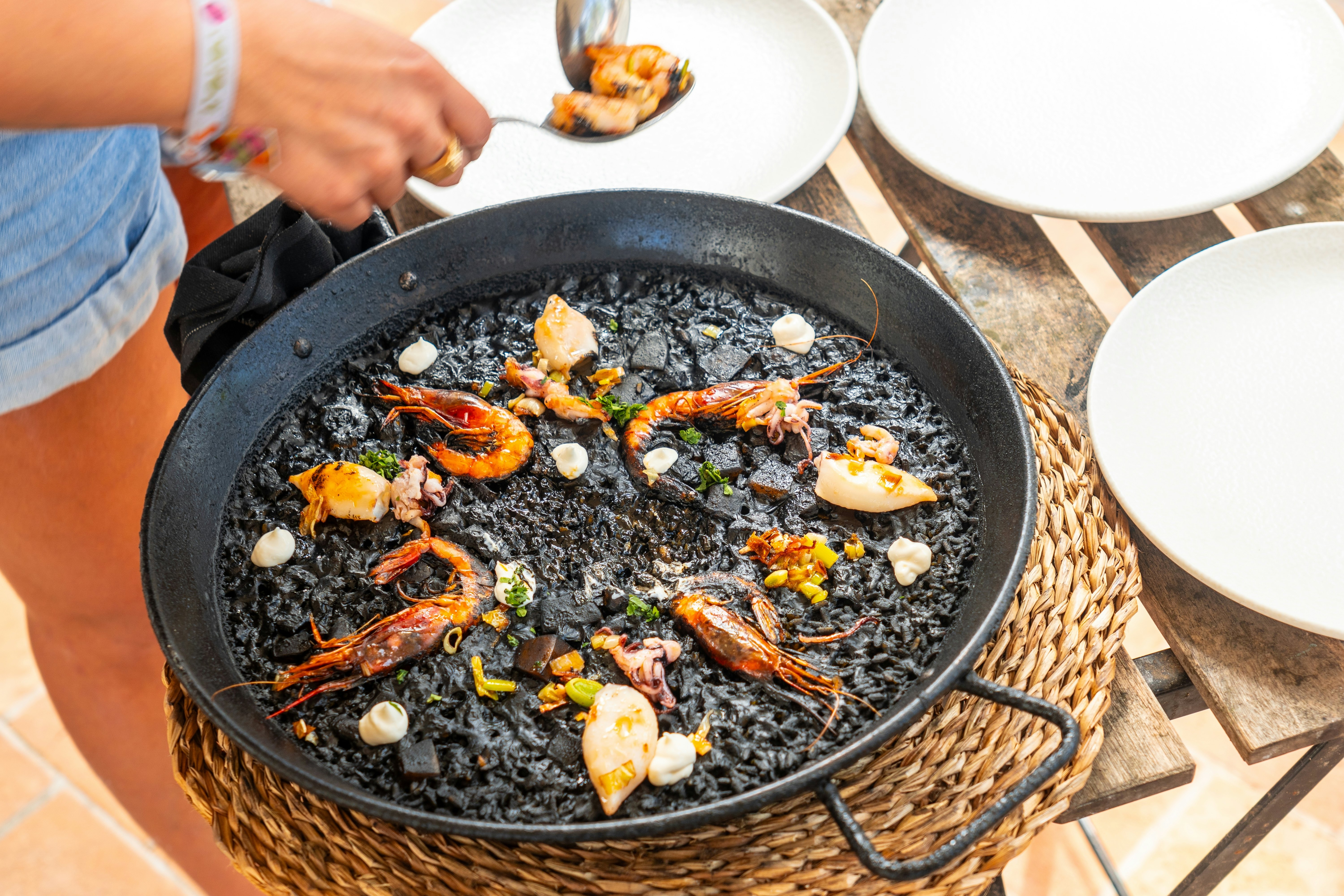Chef serving a traditional black rice paella with shrimps and seafood, using a spoon, directly from the pan, in mallorca, License Type: media, Download Time: 2025-02-03T19:04:53.000Z, User: katelyn.perry_lonelyplanet, Editorial: false, purchase_order: 65050 - Digital Destinations and Articles, job: WIP, client: WIP, other: Katelyn Perry