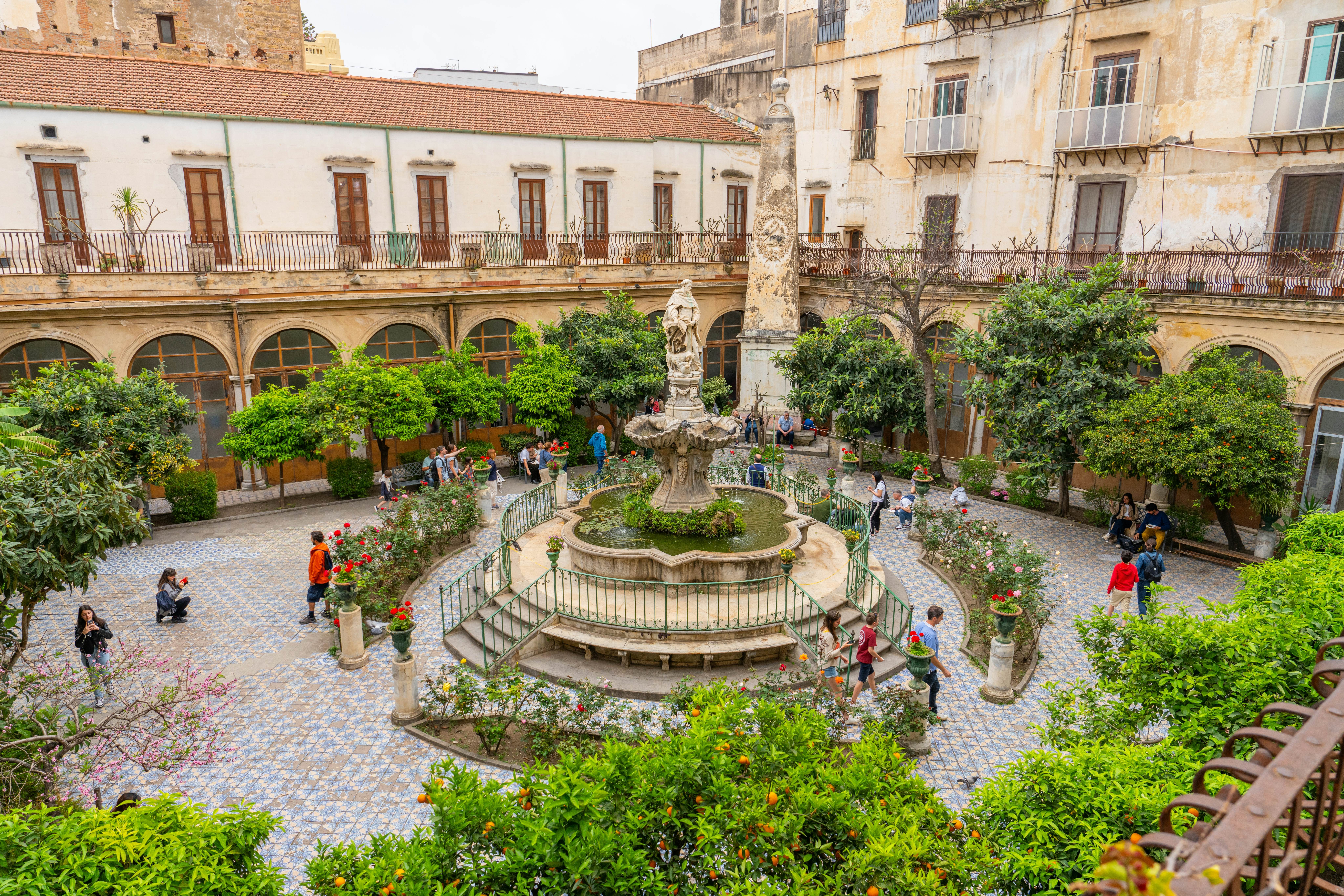 People are seen in the large courtyard of a building. Leafy orange trees surround the perimeter of the courtyard, an a sculptural fountain stands at its center.