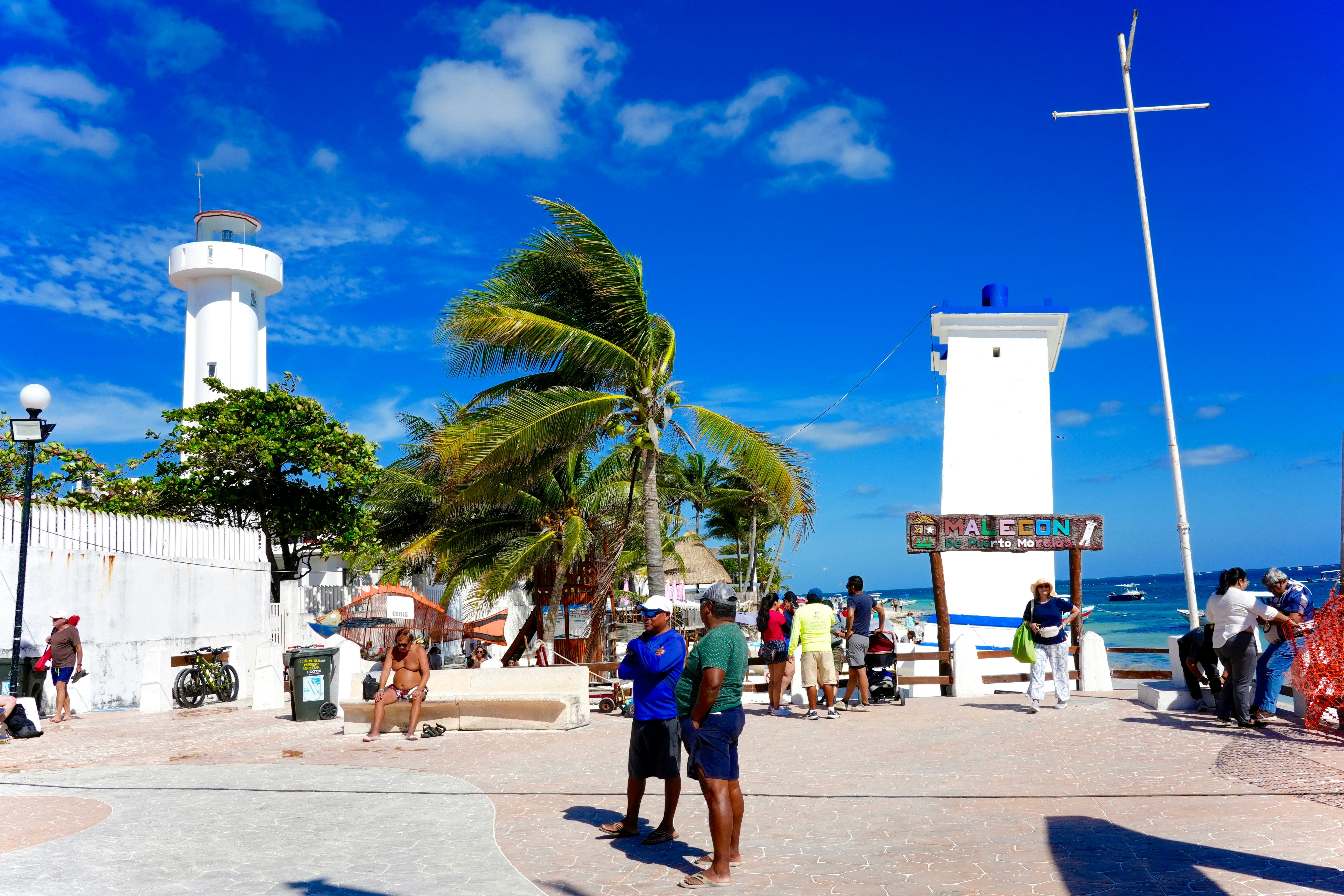 Men in shorts stand in a plaza in a village by the sea. Two lighthouses and palm trees are visible in the background.