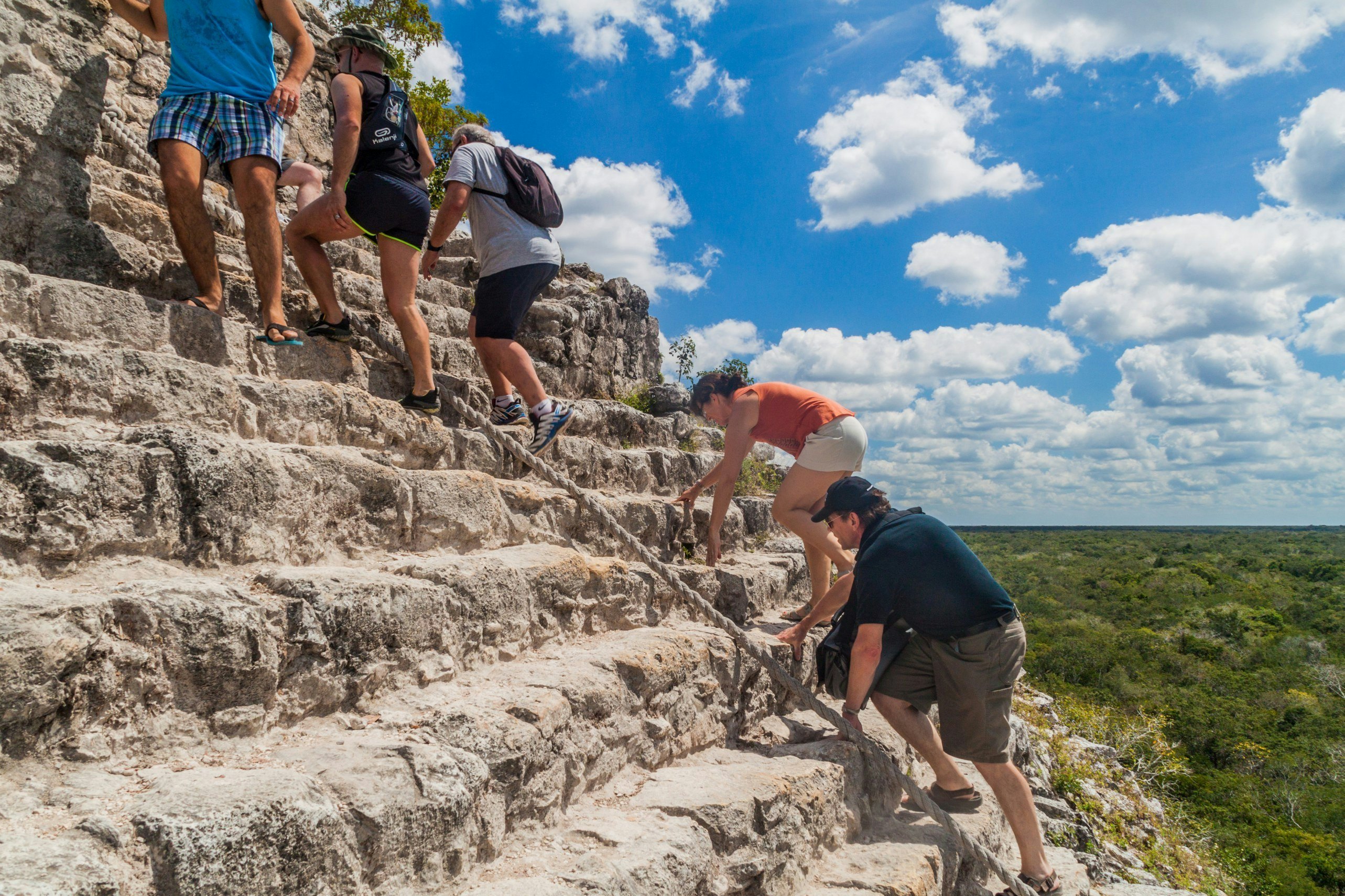 People use a rope to steady themselves as they climb a stepped pyramid. Green trees are visible below.