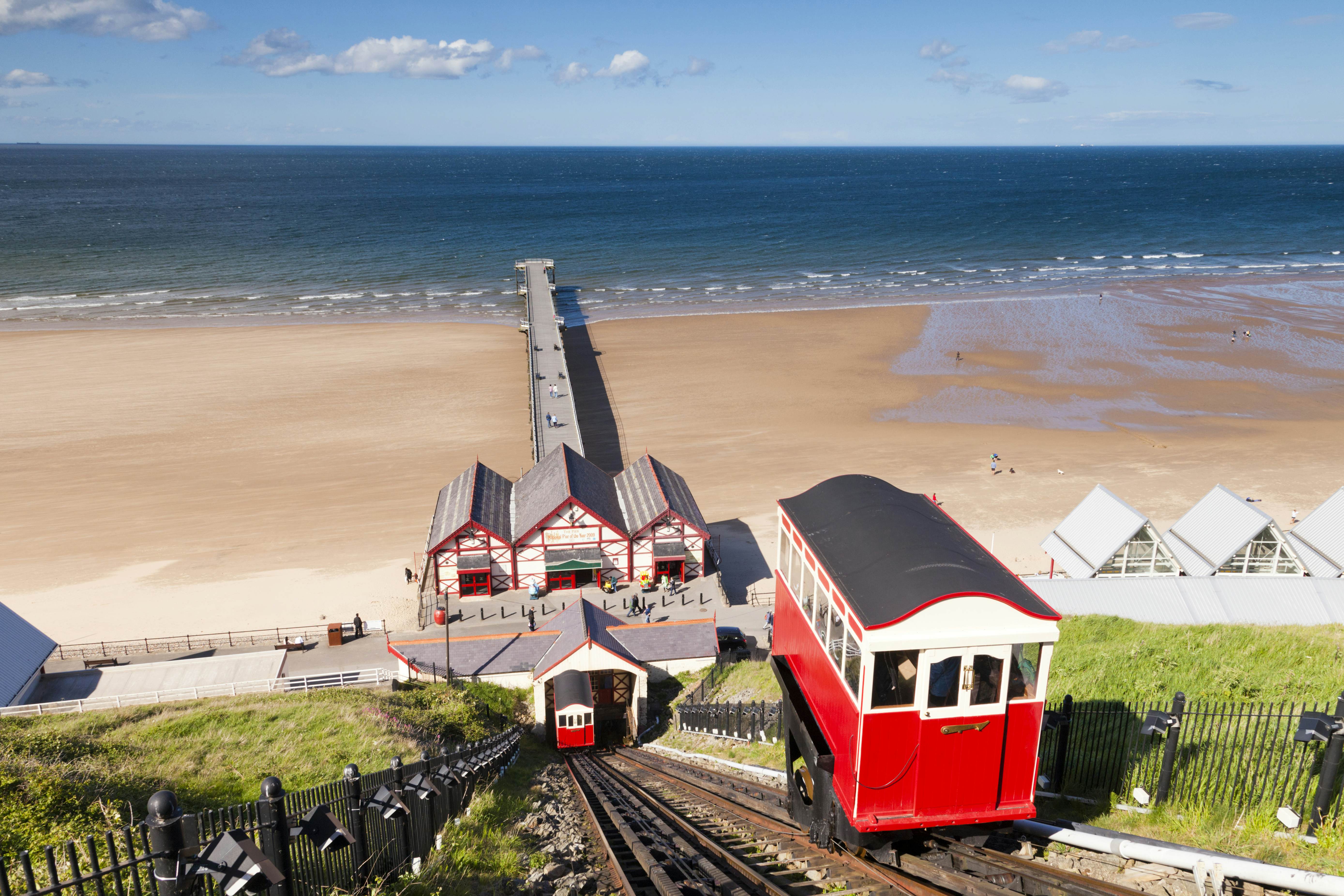 A small red funicular carriage travels down the track towards the beach and pier as its opposite carriage comes up