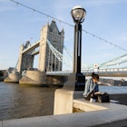 A woman reads a book on a ledge, with Tower Bridge behind her.