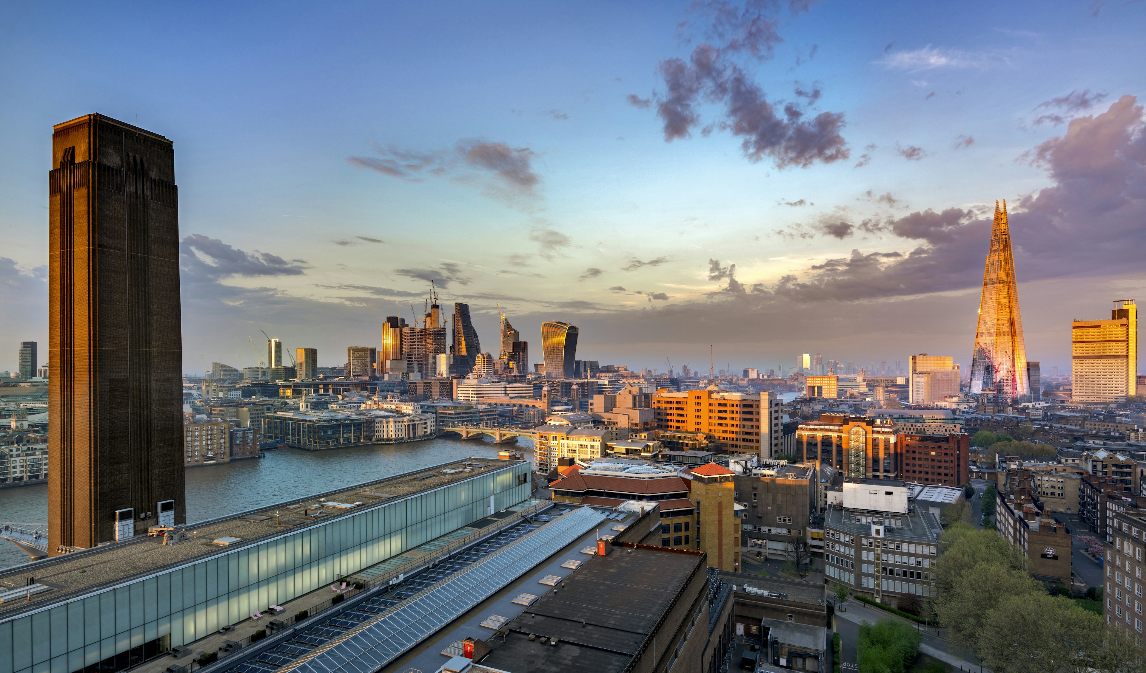 A city view including a large industrial building with a tall chimney contrasting with a pointed glass and steel skyscraper