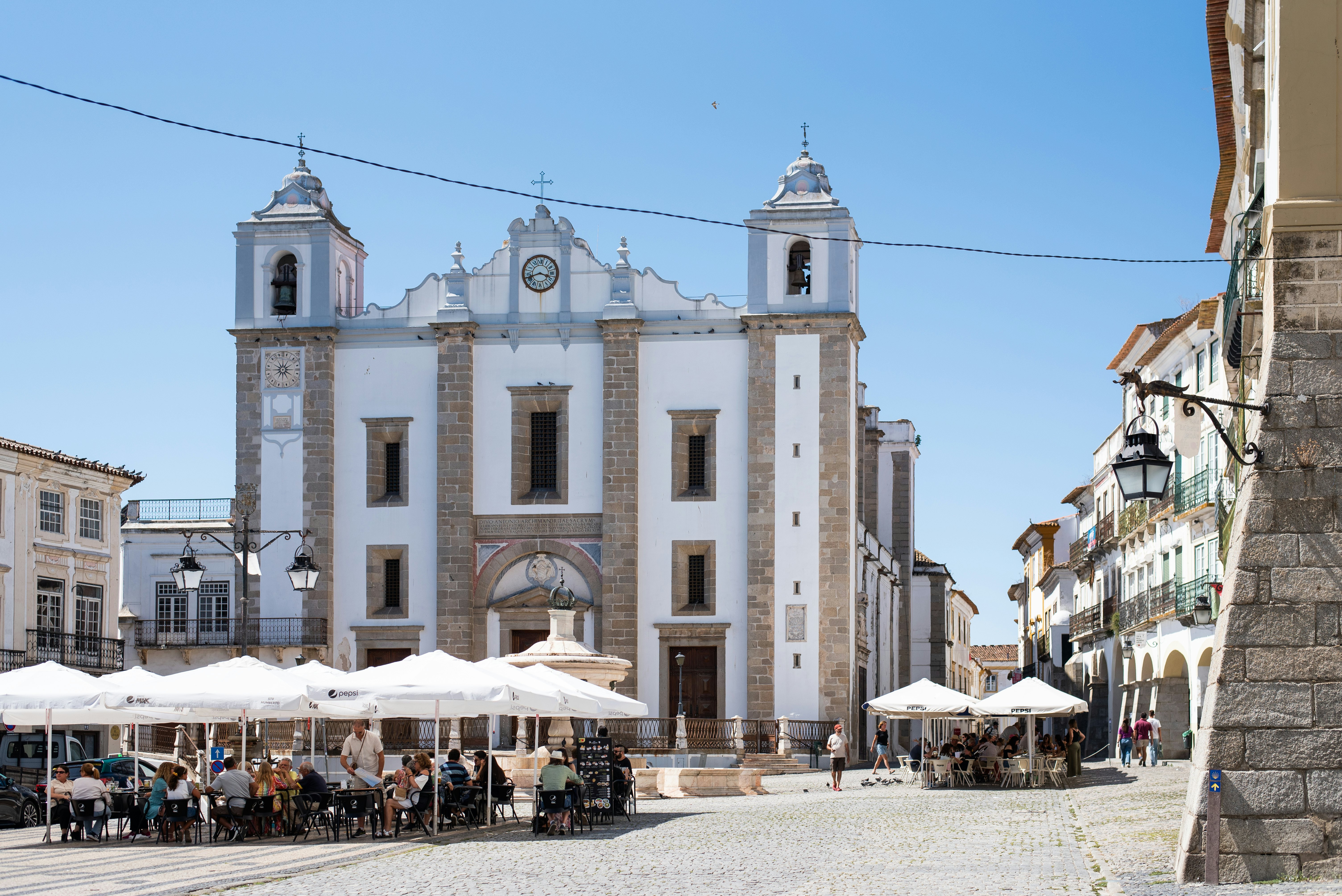 People sit under umbrellas near white stone buildings in a public square.