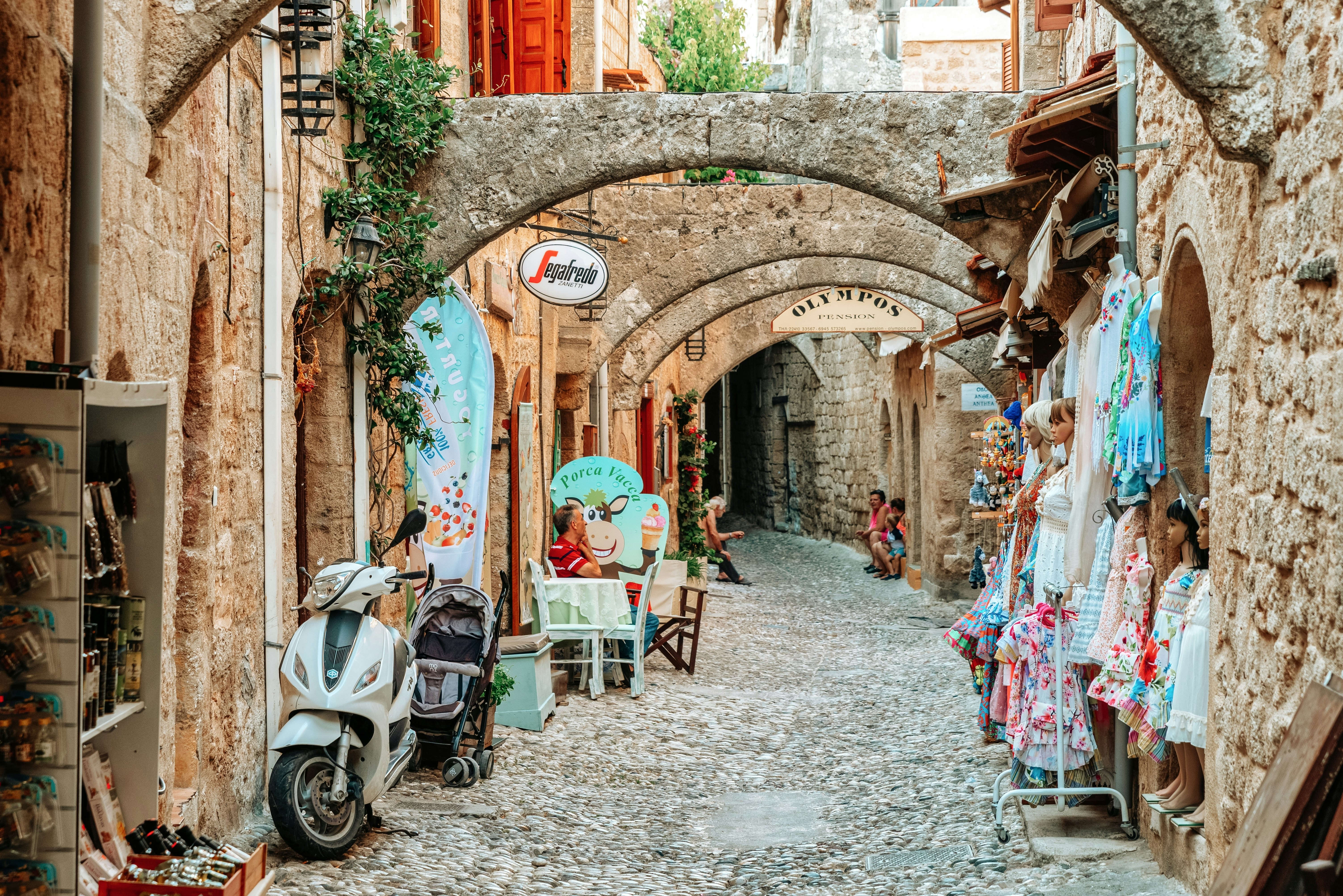 A narrow, cobbled street in a historic town is lined with shops, and covered with arch supports