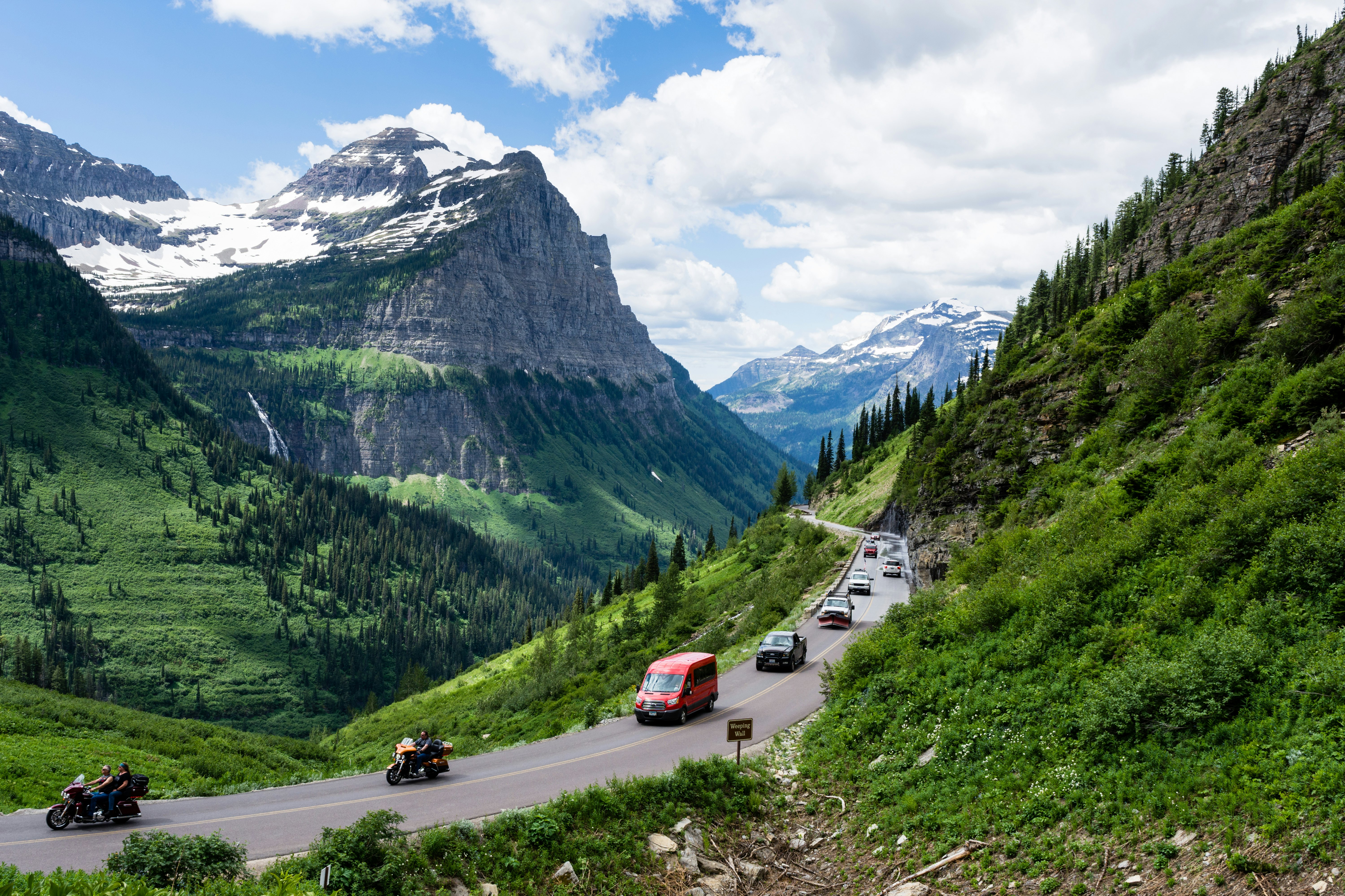 A line of cars drives along green grass and a mountain road