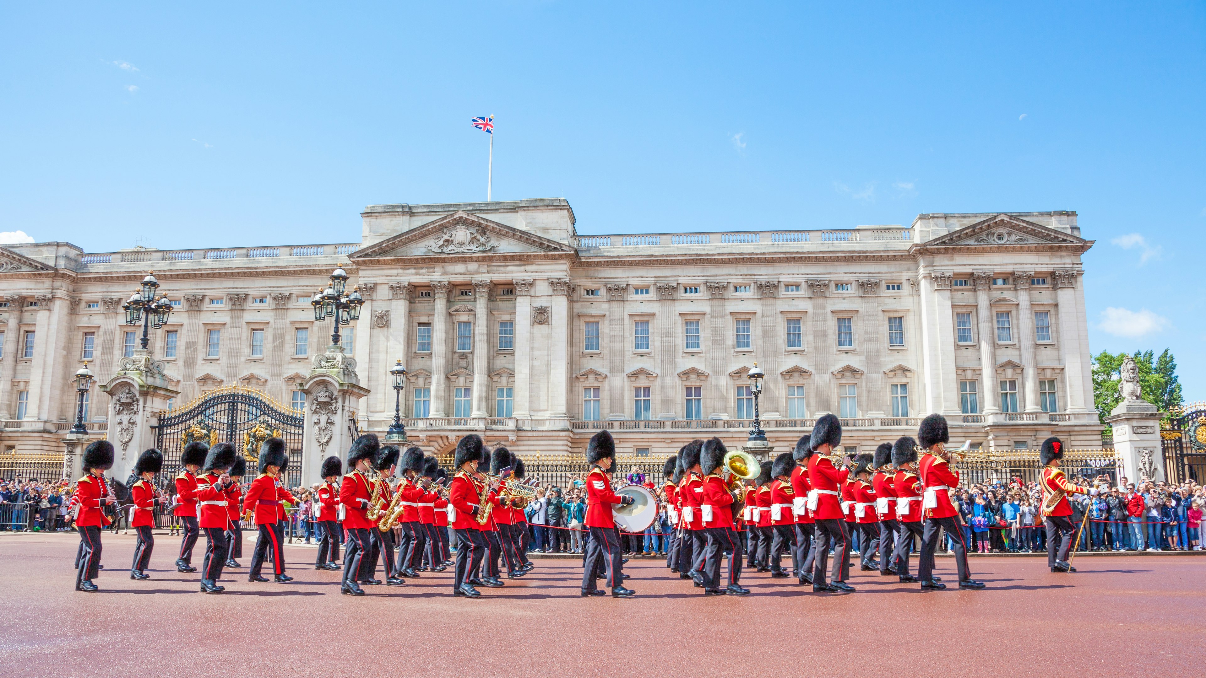 Guards wearing red uniforms and bearskin hats march in front of a crowd outside a vast palace building
