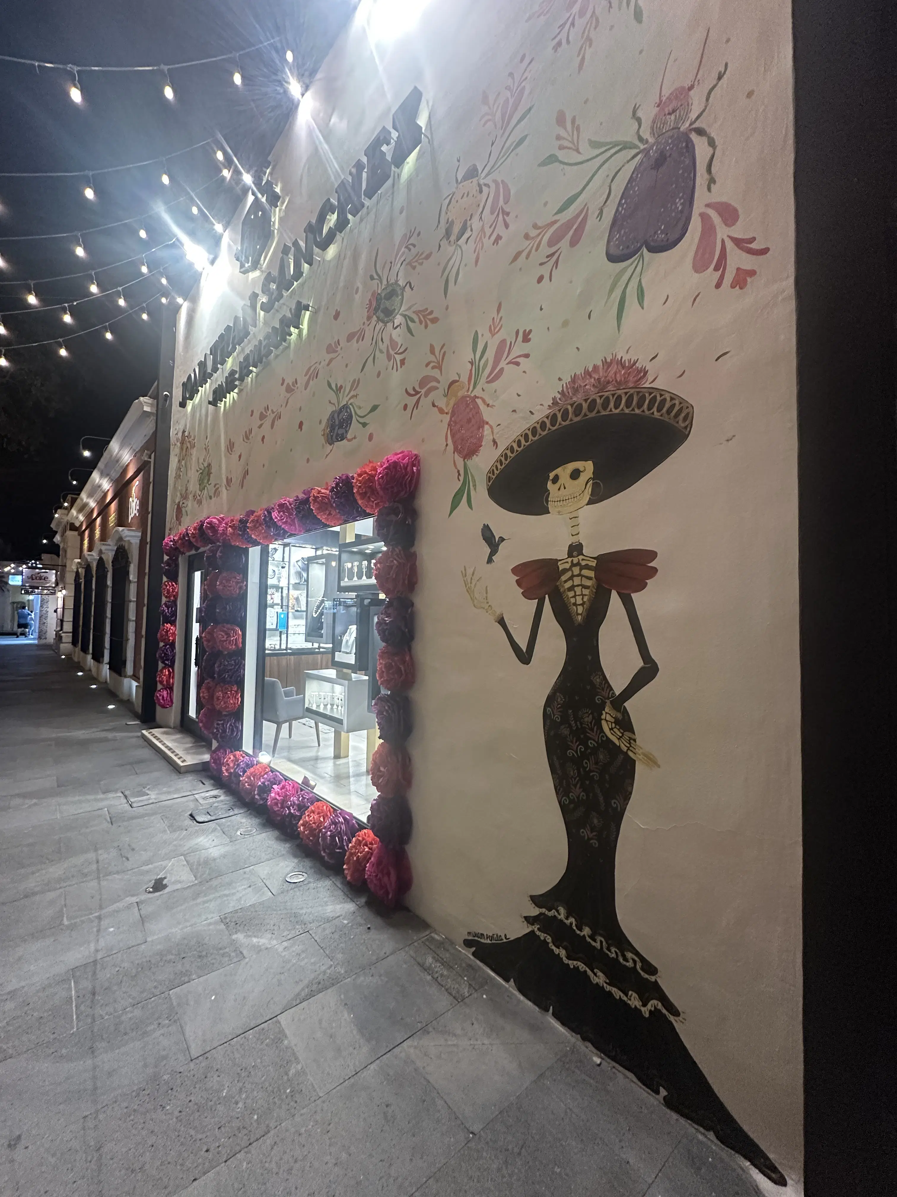 Sugar skull street art of a woman in a black dress and sombrero with pink flowers.