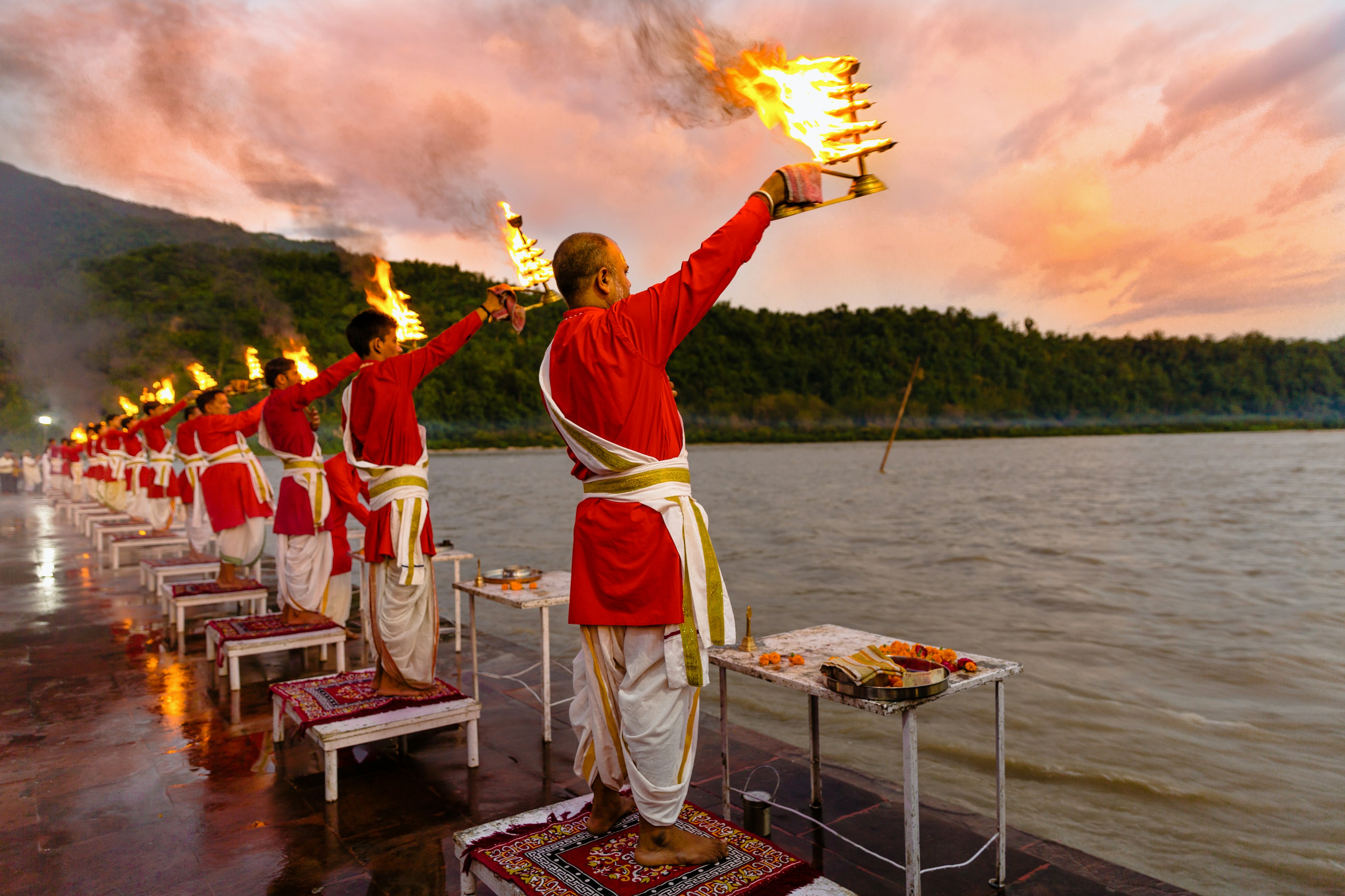 Priests wearing red tunics and white sashes raise burning lamps toward a river during a ceremony at dusk.
