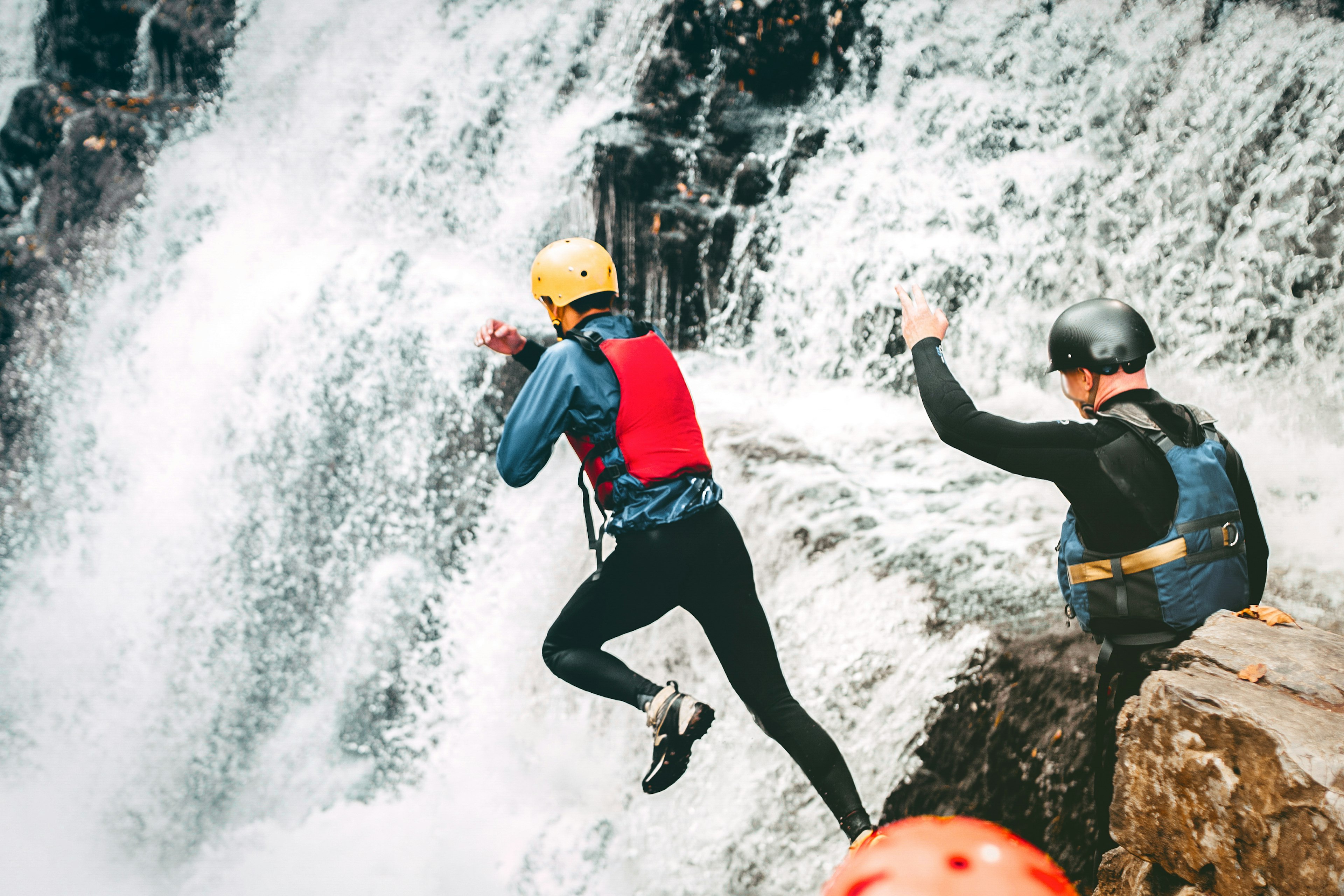 A person wearing a blue dry suit and yellow helmet jumps into a waterfall while another person wearing a black helmet looks on with his arm up.