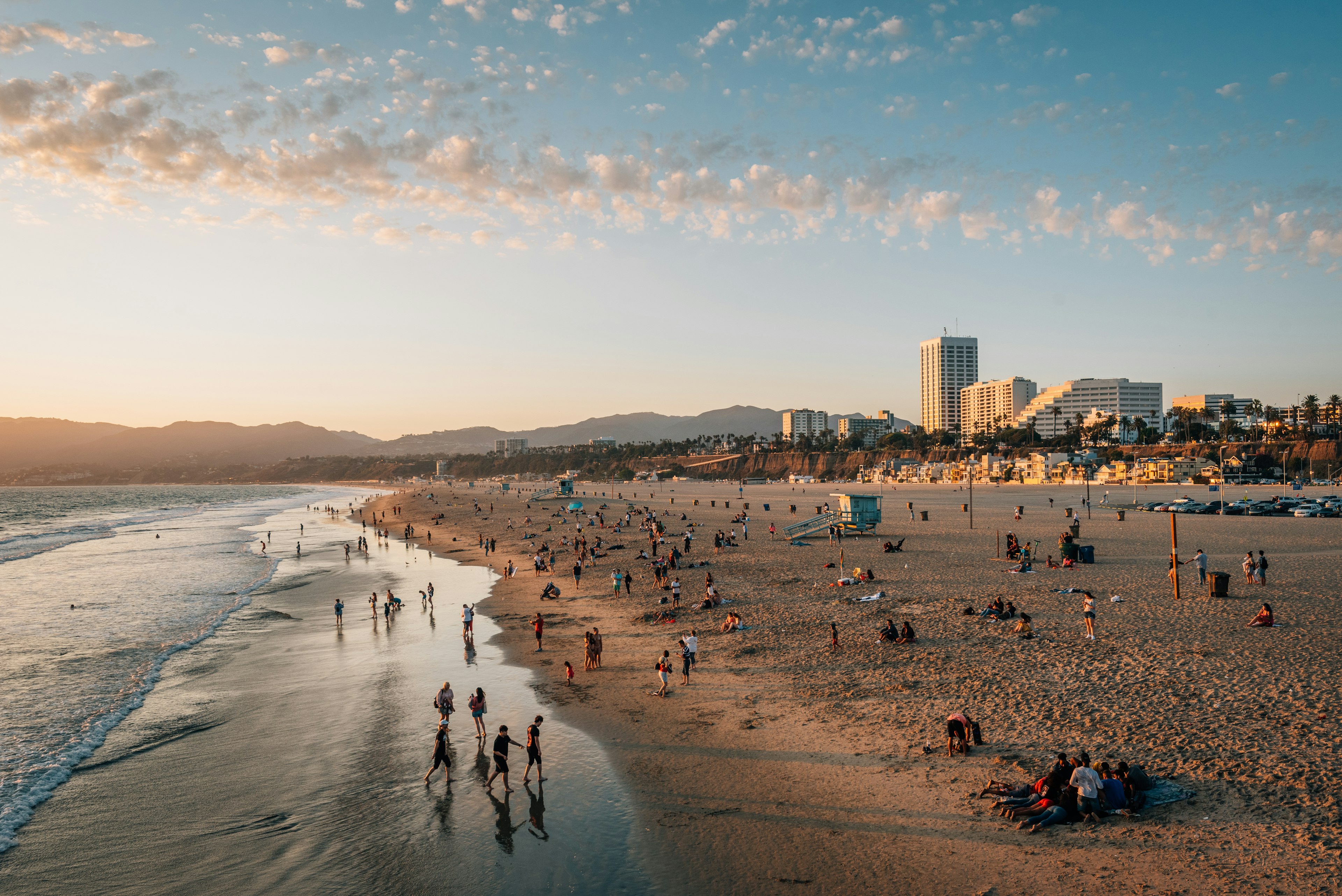 An aerial view of a beach at sunset. Numerous people sit on the beach and stand in the surf. Hills and apartment buildings are visible in the distance.