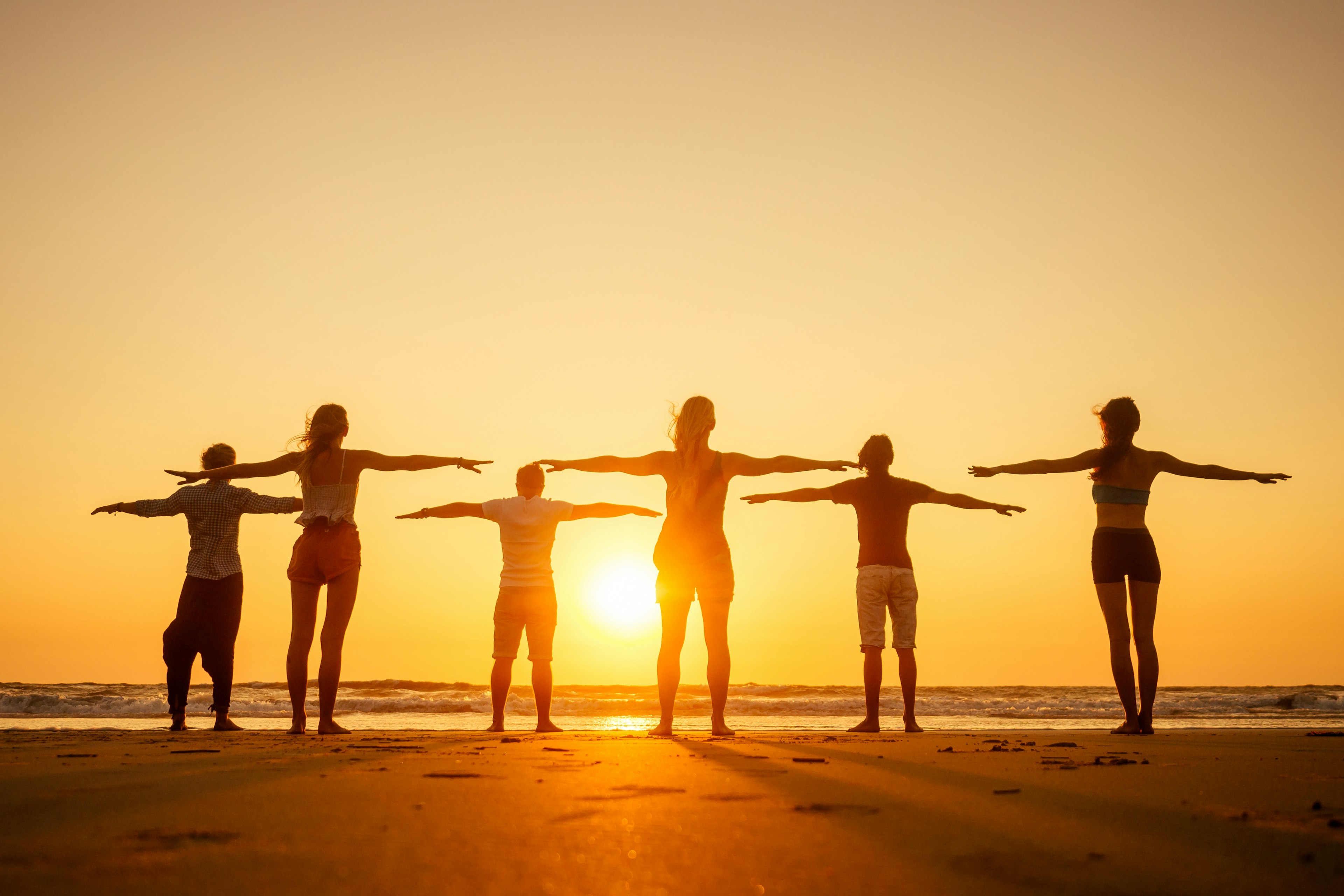 The silhouettes of six people facing the rising sun on a beach. Their arms are stretched out to the side.