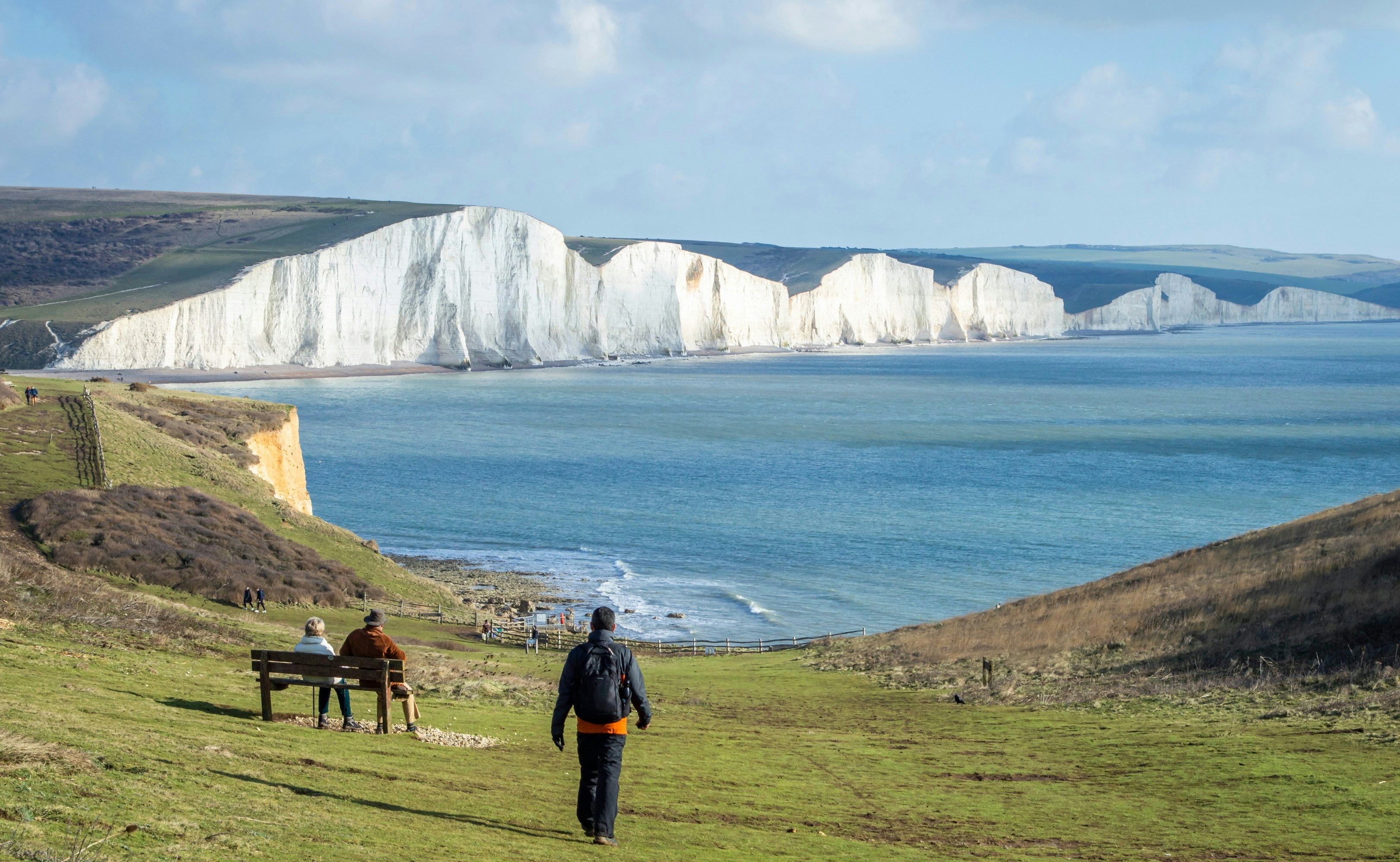 A man walking down a hillside of green grass toward a couple sitting on a bench that's facing white chalk cliffs above blue water.