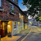 CANTERBURY, KENT, UNITED KINGDOM, SEPTEMBER 14, 2023: Warm glow of a traditional inn shines onto the street on an early summer evening. A charming brick house facade with flower boxes in Canterbury., License Type: media, Download Time: 2025-04-08T14:26:10.000Z, User: tasminwaby56, Editorial: true, purchase_order: 65050 - Digital Destinations and Articles, job: Online Editorial, client: Canterbury TTD, other: Tasmin Waby