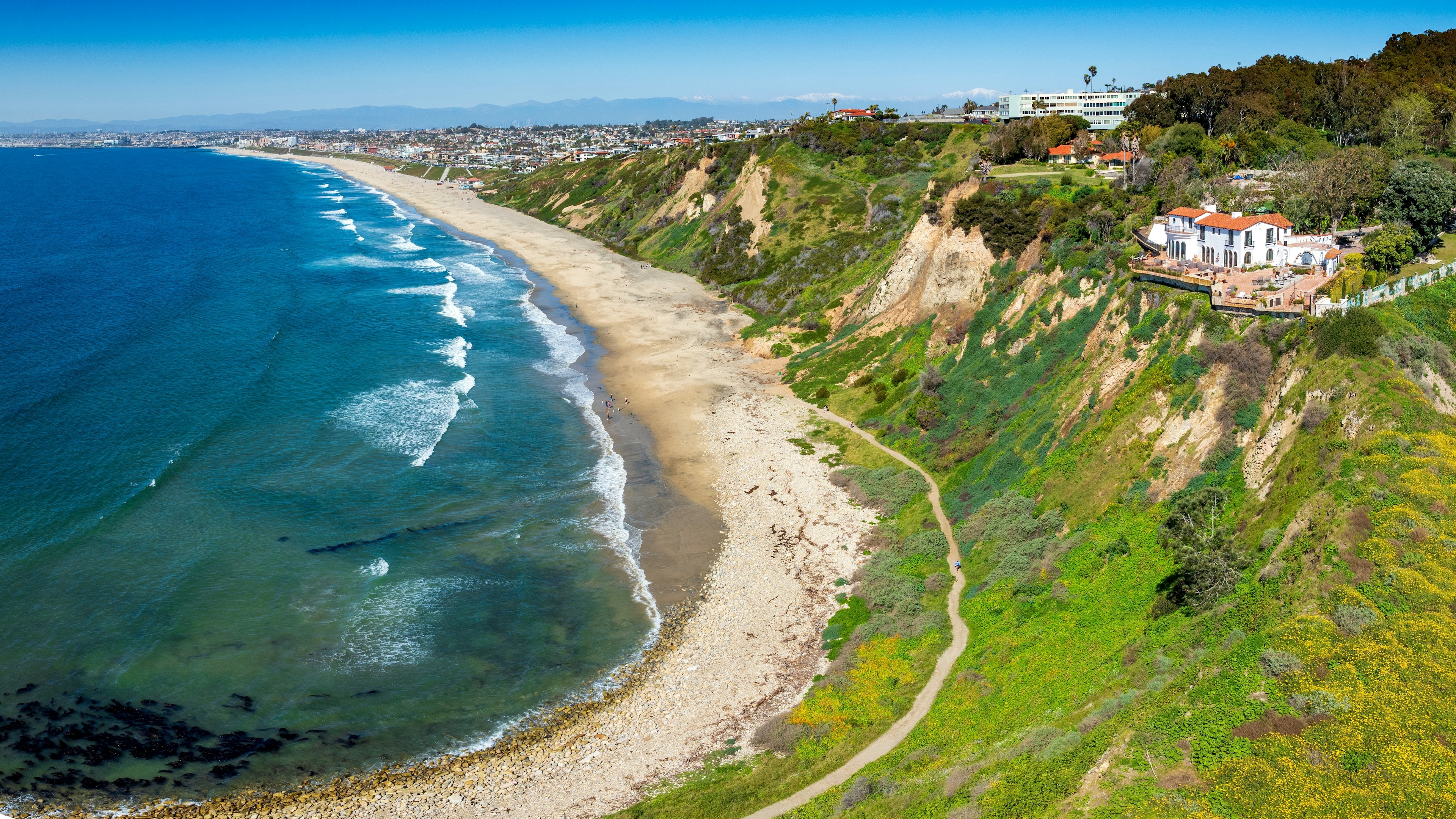 Aerial view of curved coastline beach with ocean to the left, homes on a hillside to the right, and a city view in the distance.