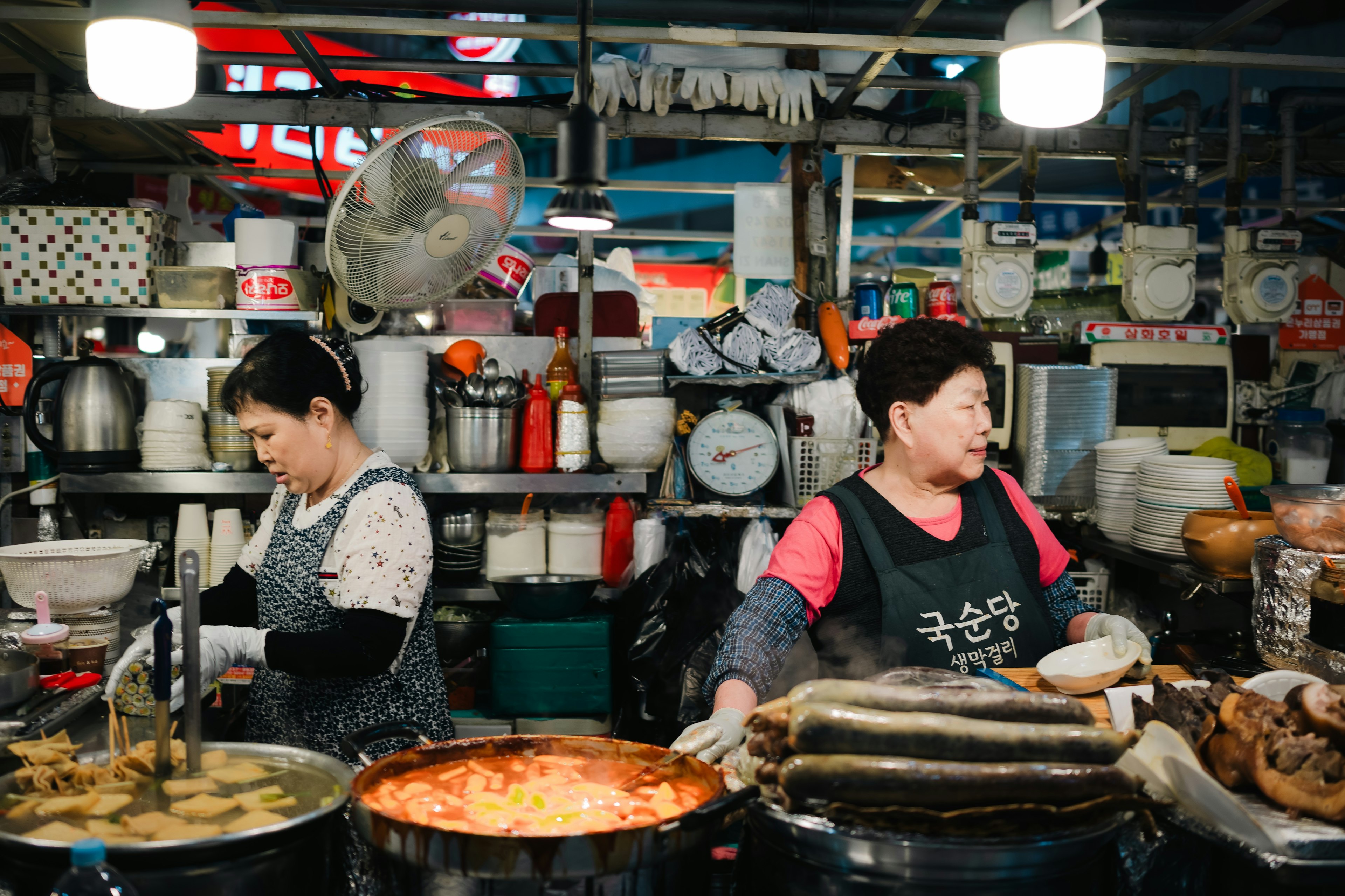 Two women prepare large pots of food in a small kitchen stacked with supplies