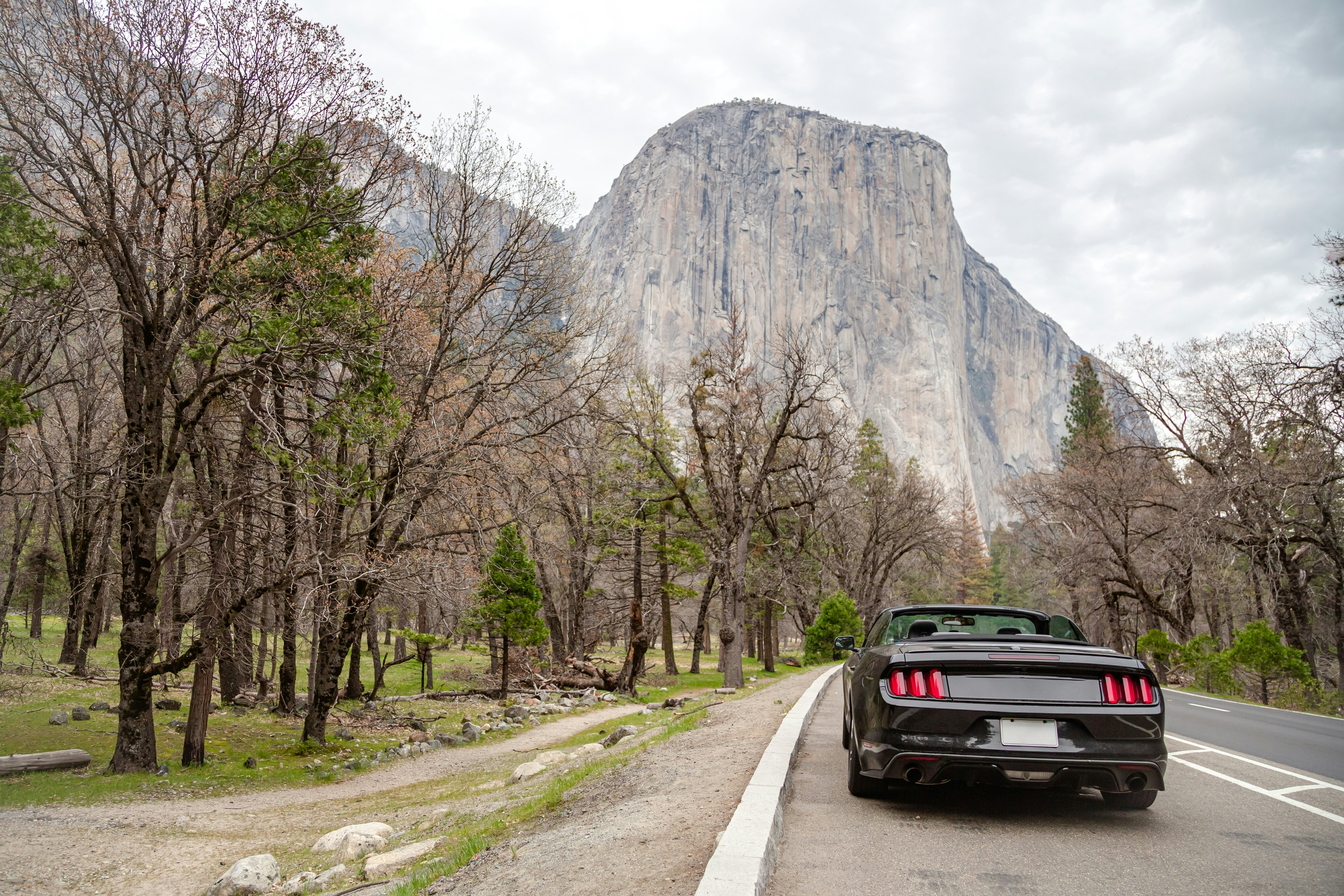 A black convertible drives on a road through a park. A huge rock formation is visible in the distance.