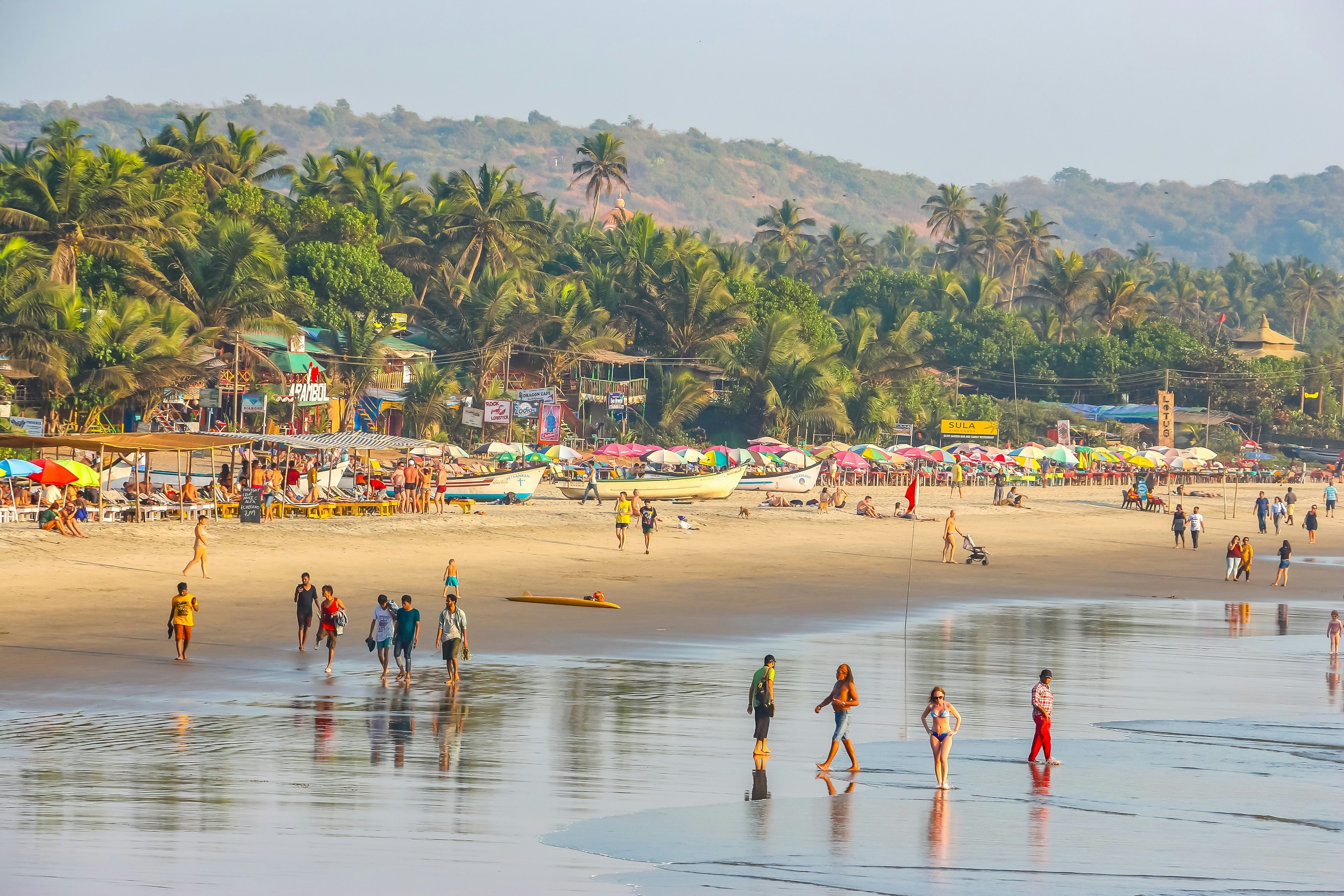 A wide view of people walking in the surf of a wide tropical beach. Palm trees line the shore beyond boats and shacks on the beach.