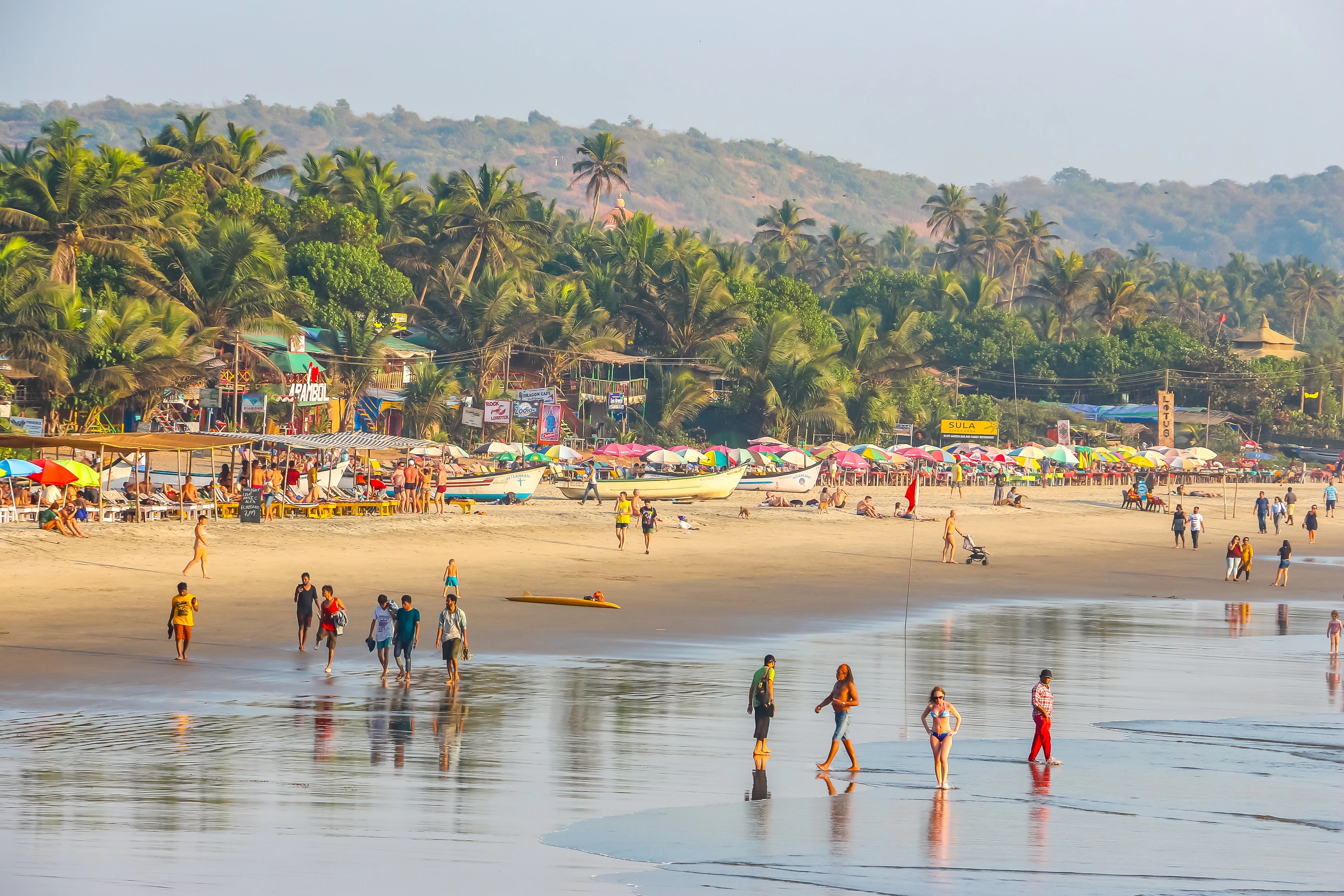 A wide view of people walking in the surf of a wide tropical beach. Palm trees line the shore beyond boats and shacks on the beach.