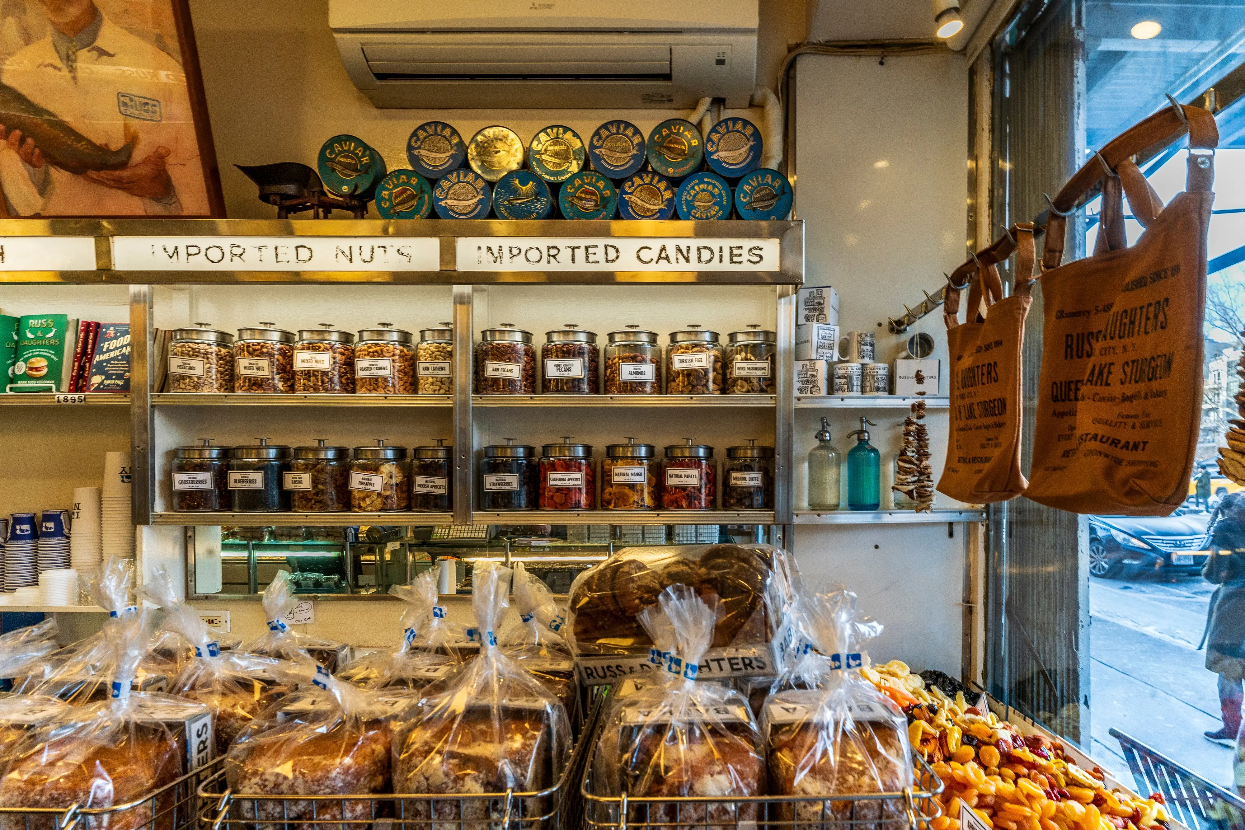 Russ and Daughters, a historic NYC appetizing shop with shelves displaying nuts, candies, and caviar. Baked goods, dried fruits, and branded tote bags are visible.