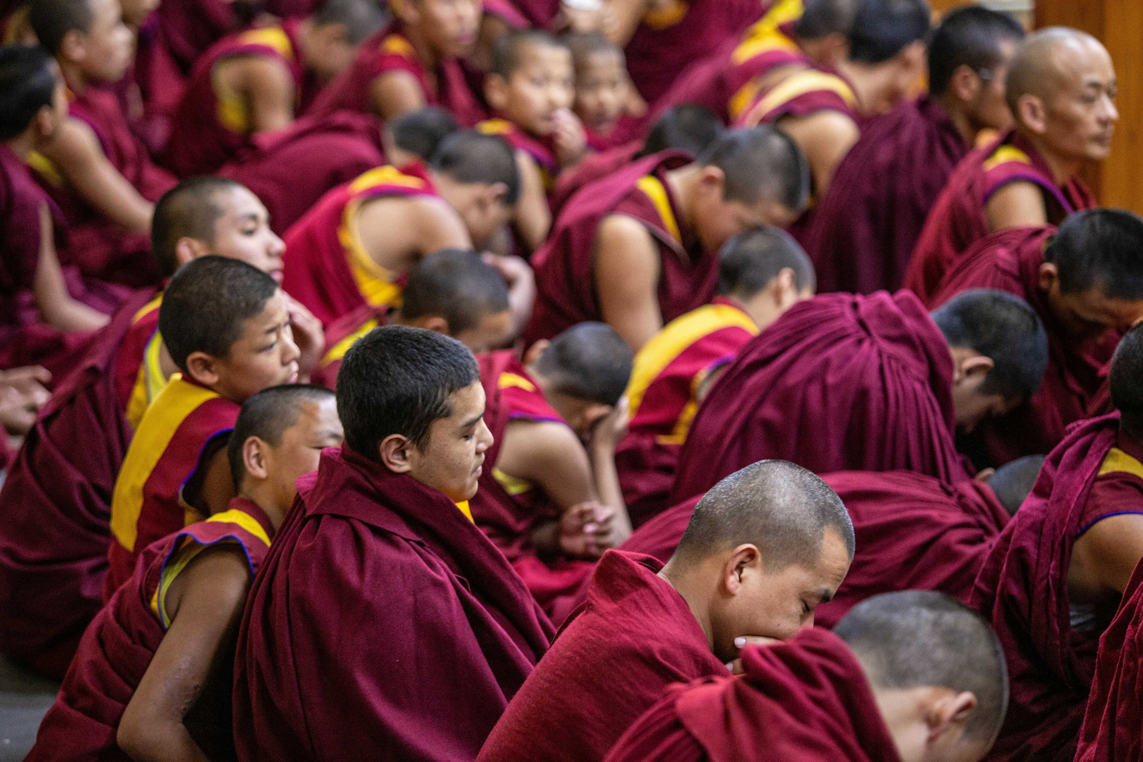 Monks in burgundy robes with yellow sashes sit with their heads bowed.