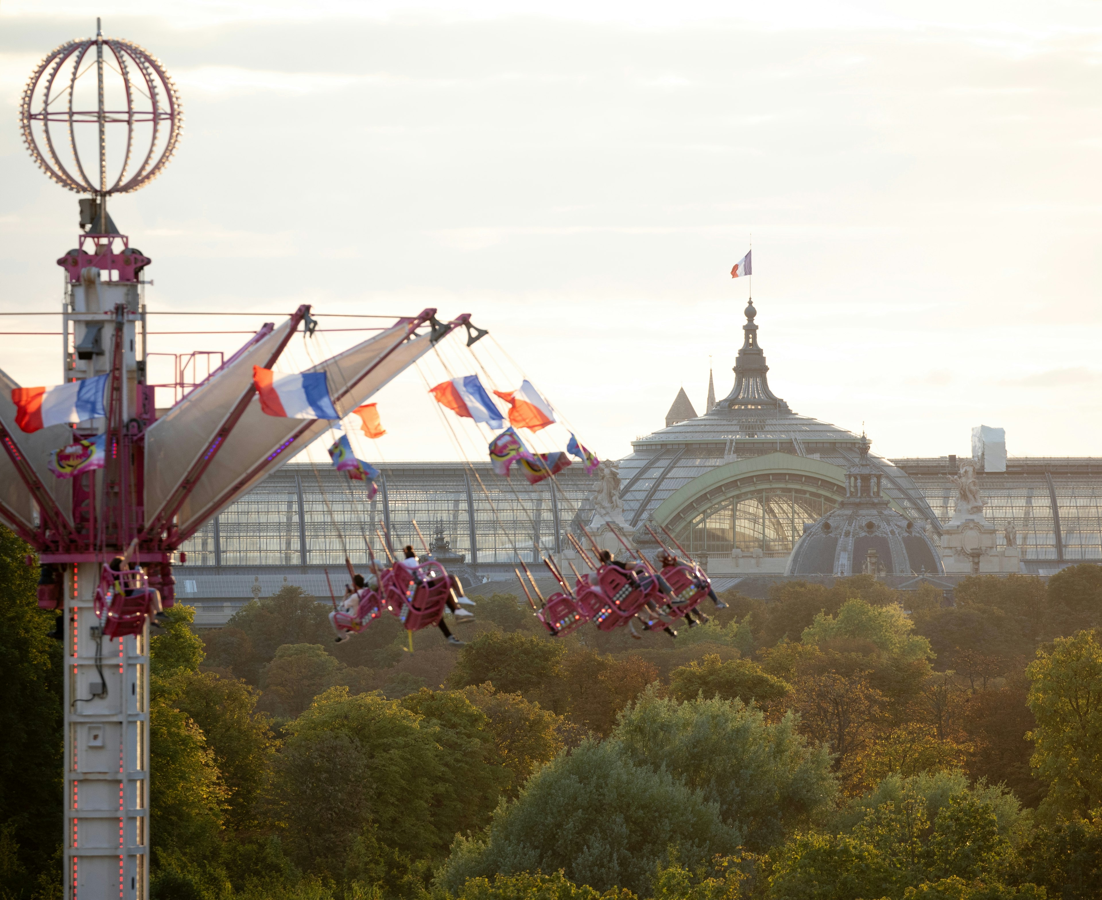 Carousel of flying swings spins over green trees with Grand Palais in Paris in the background