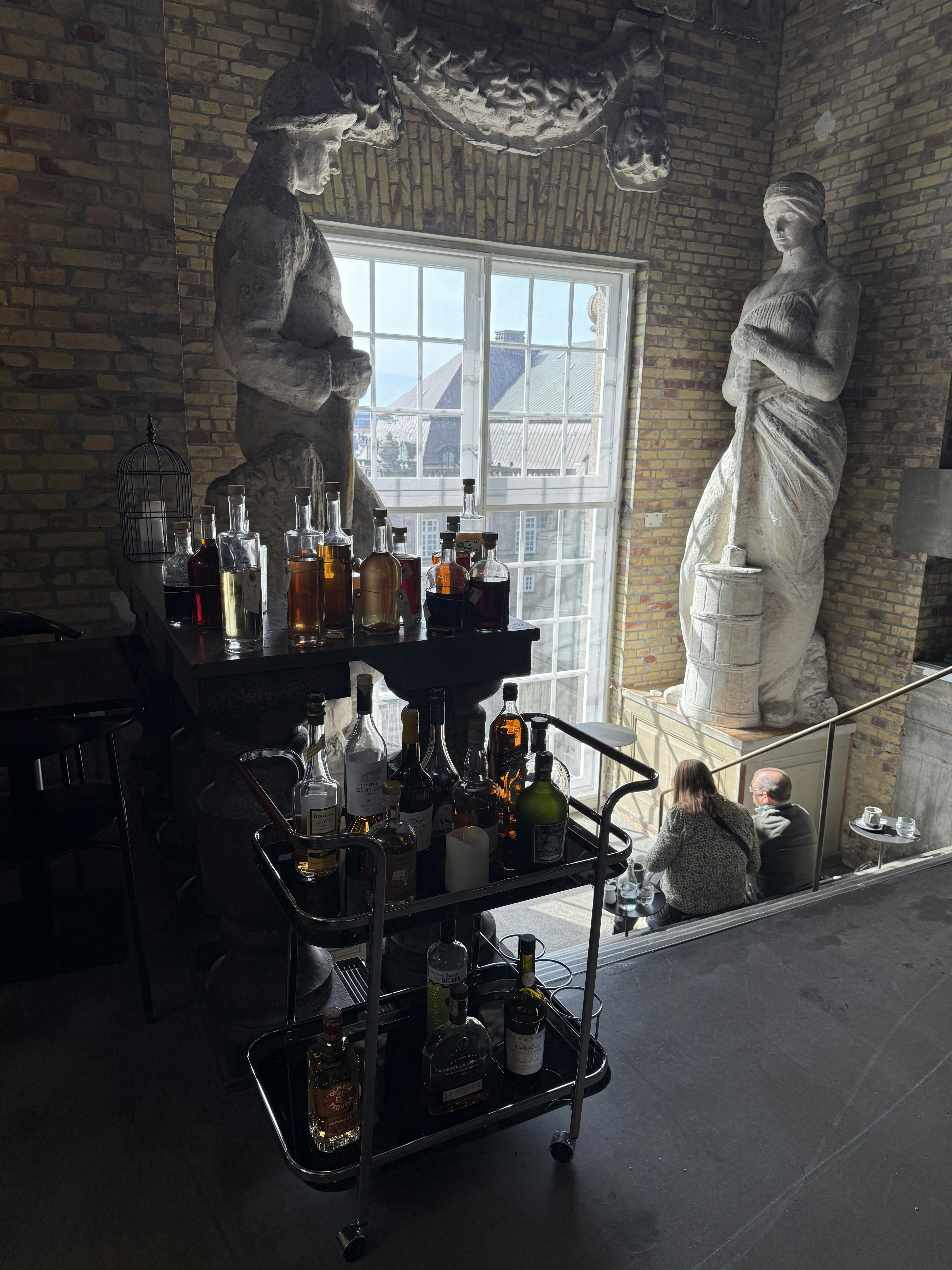 Close-up of bar and rolling tray containing bottles of alcohol with tall, white sculptures of people surrounding a tall window and a couple sitting on cement stairs.Inside the tower at Tårnet in Cophenhagen