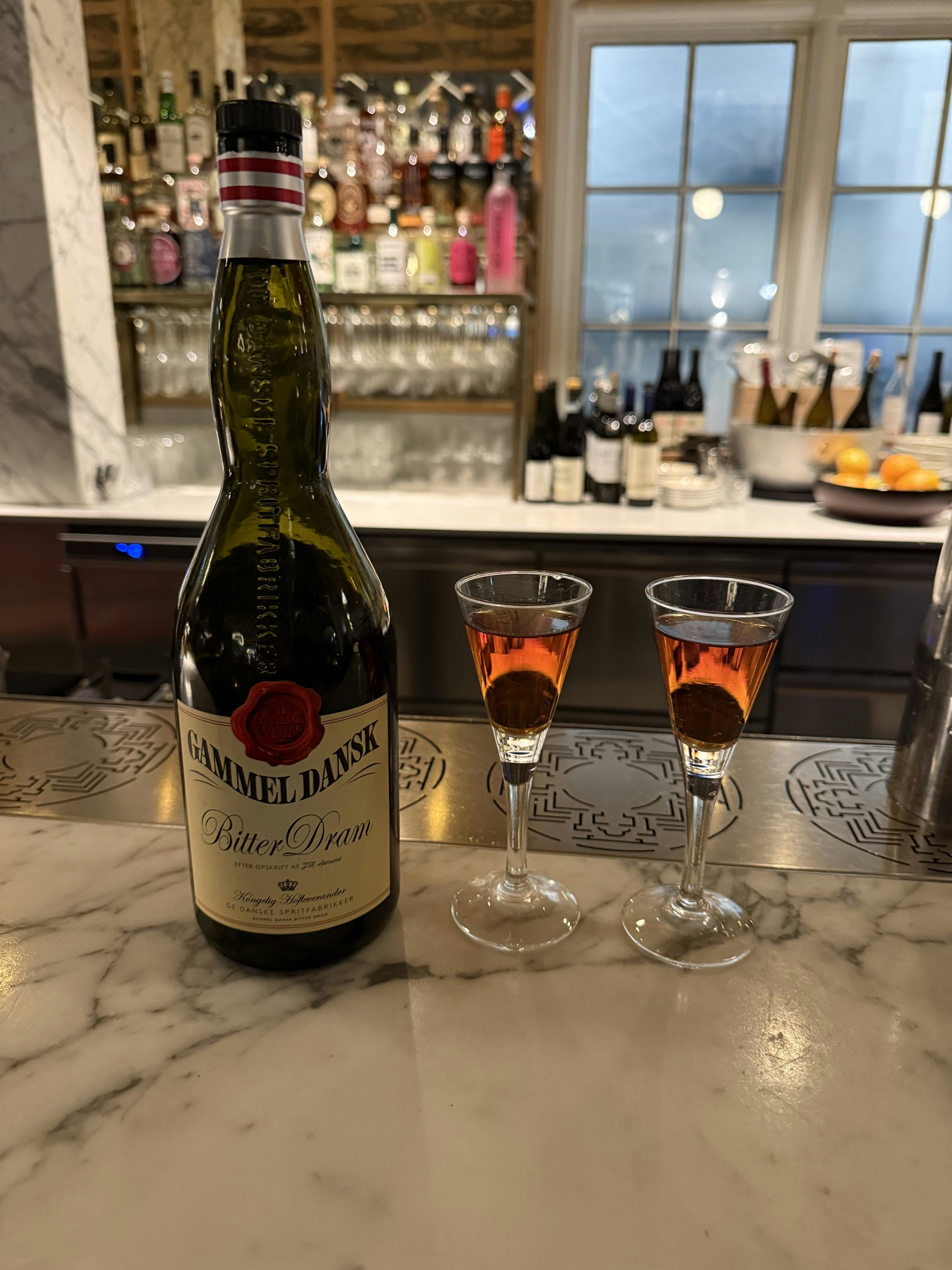 Close-up of a Gammel Dansk bottle on a white marble bartop with two fluted glasses to the right filled with browinsh liquor.
