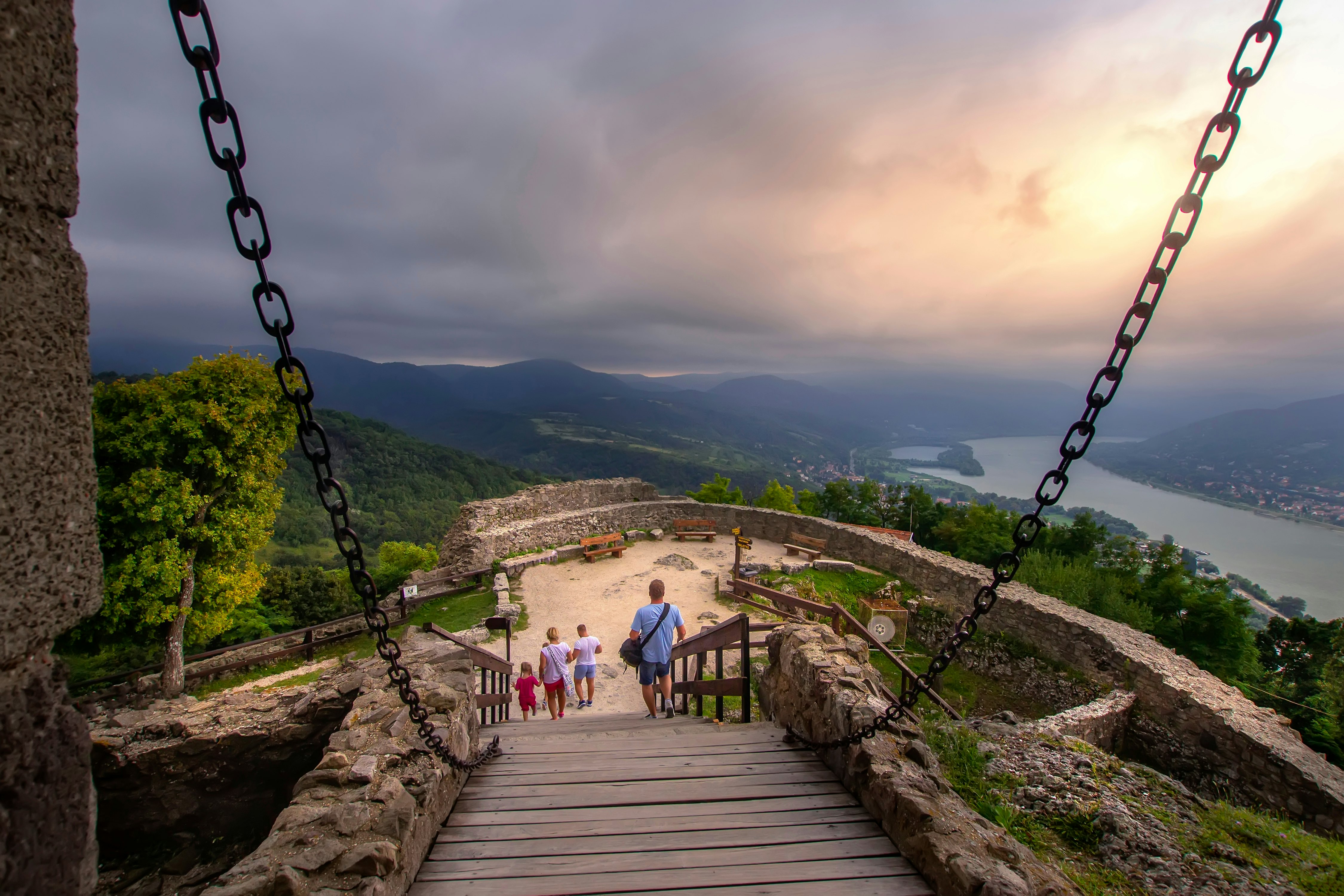 People walk down a ramp toward an overlook far above a river; a stone wall surrounds the overlook, and there are low stone walls leading to the ramp, with chains connected that rise to a point out of view.