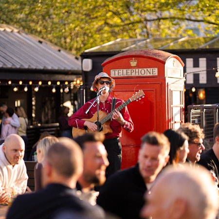 A man in a bucket hat plays the guitar and sings in a pub beer garden.