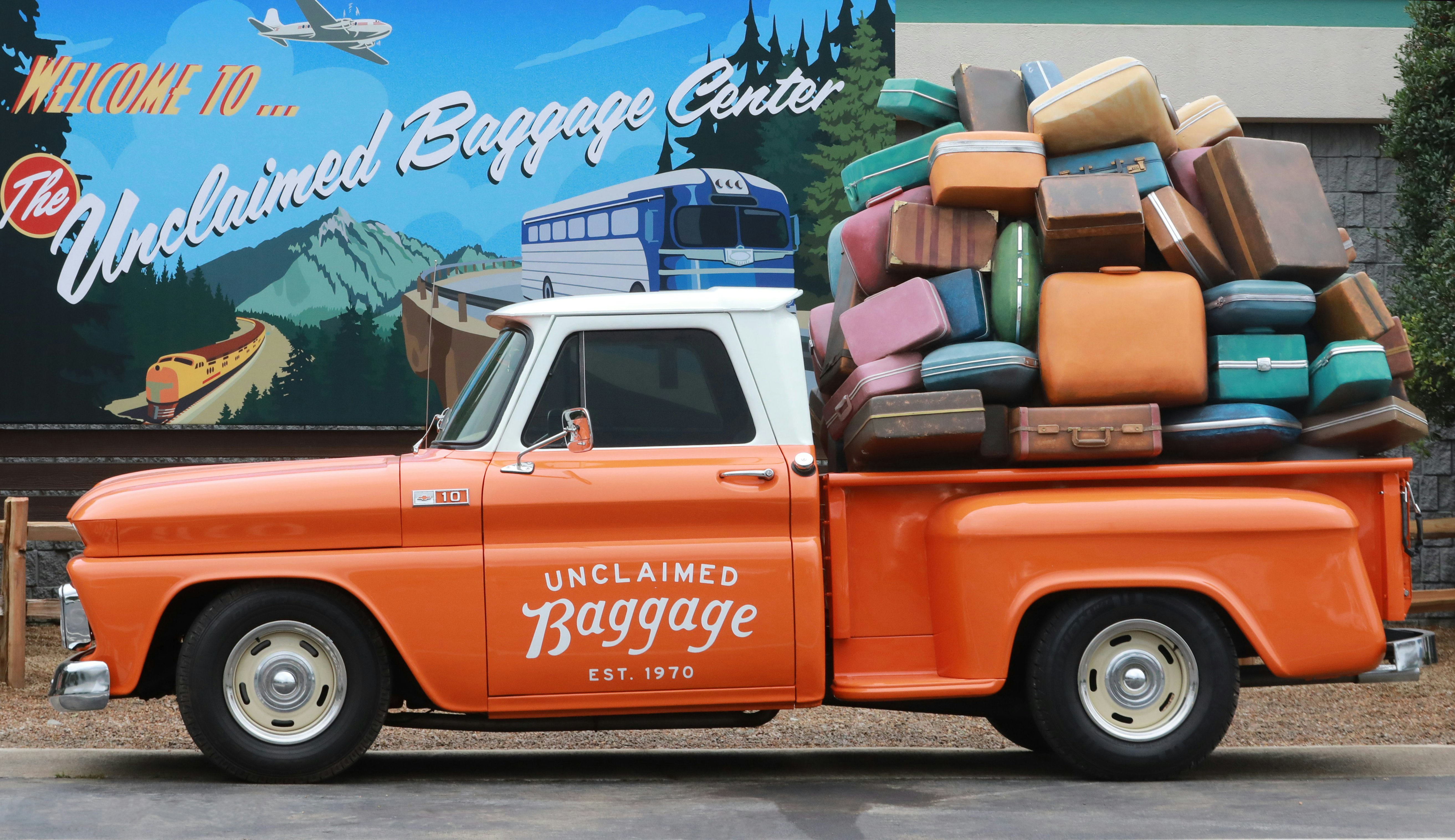 An orange pickup truck filled with suitcases, labeled "Unclaimed Baggage"