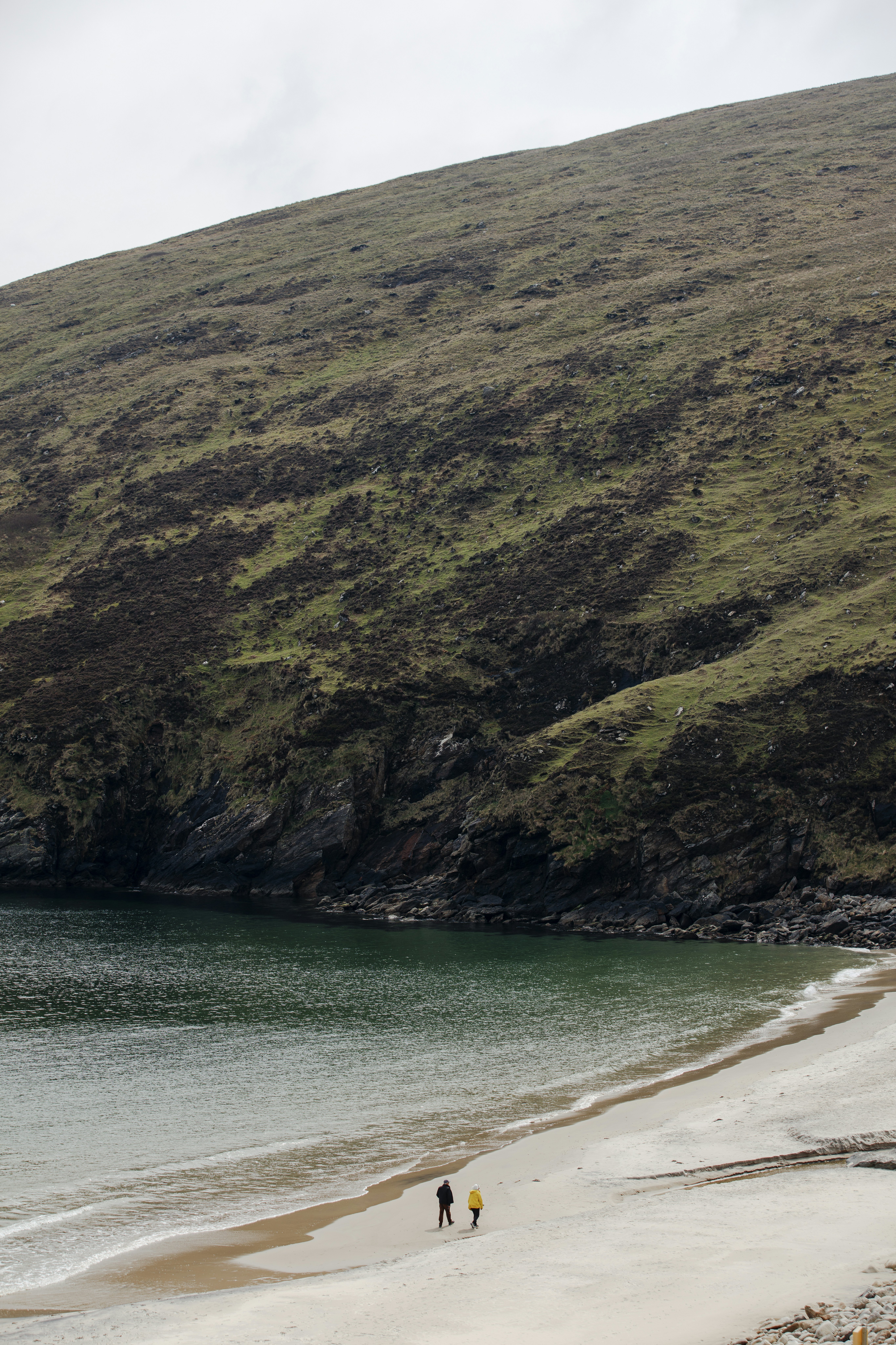 Wide shot of couple in winter gear walking on wide sandy beach with the sea to the left and an imposing, grass-covered tall hill in front of them.