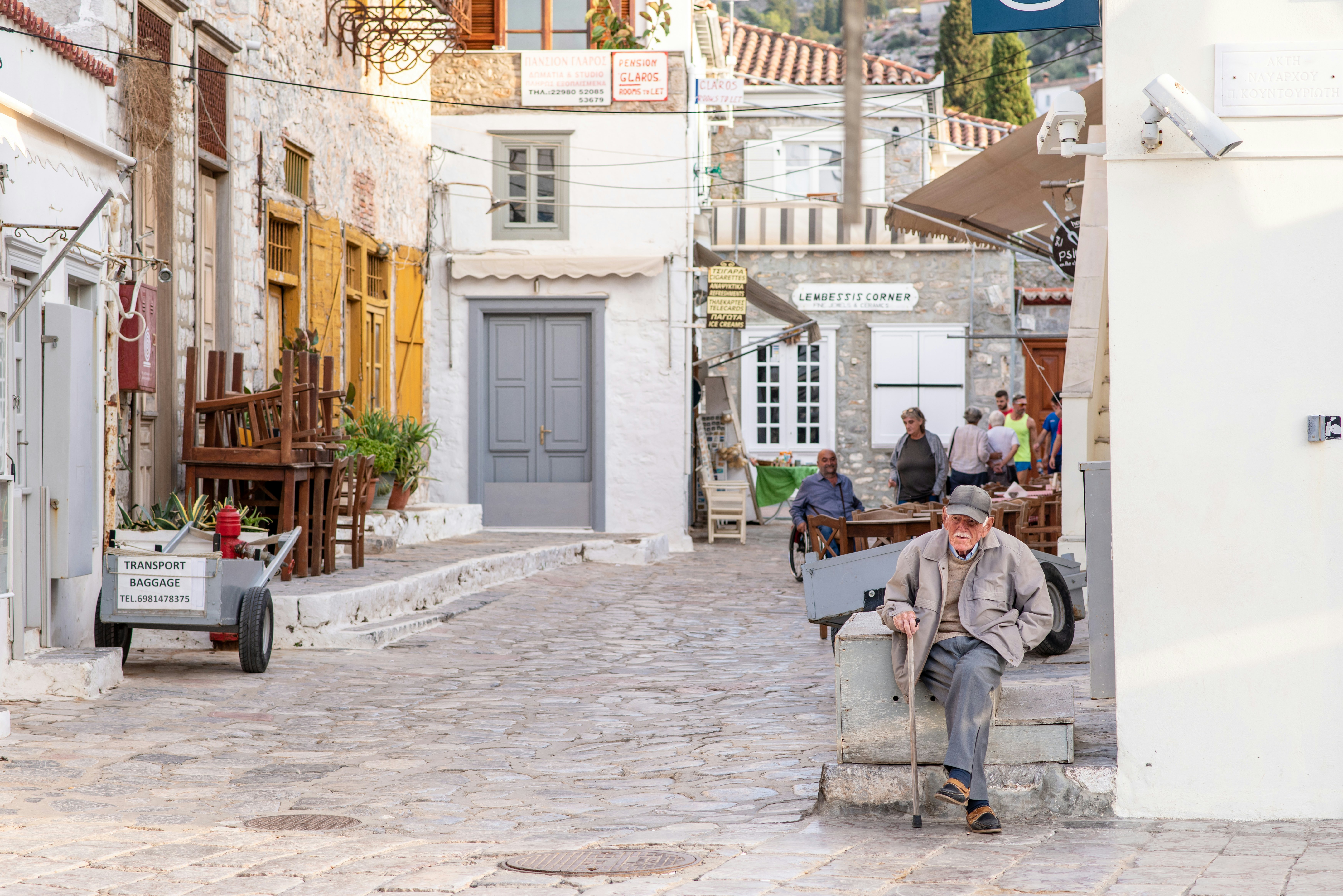 An older man with a cane sits on a wooden step with his legs crossed. Old buildings with some splashes of color on their windows and doors are in the background.