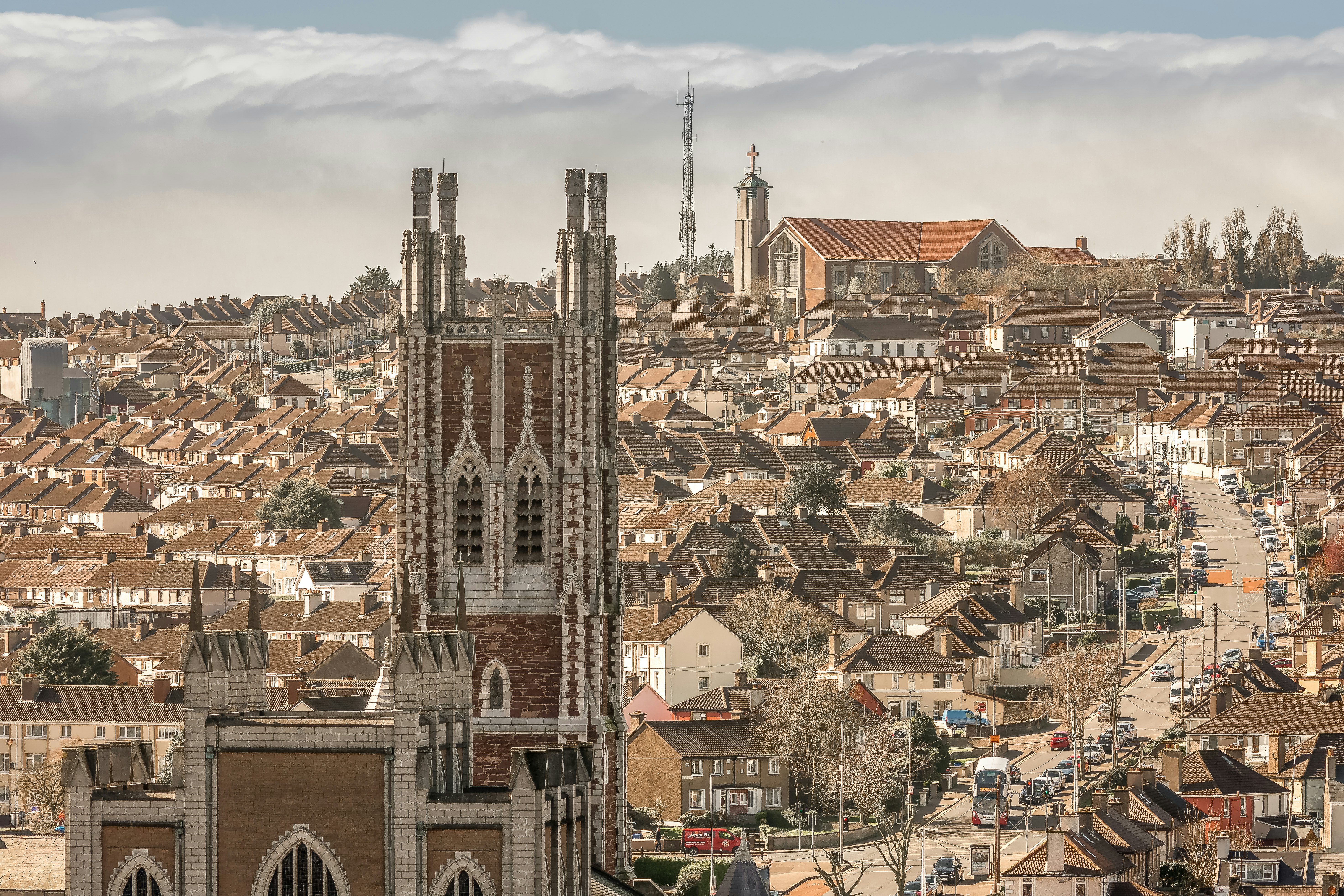 A church with a tower is seen in the foreground of a city scene, with dense clusters of houses rising on a hill behind the church.