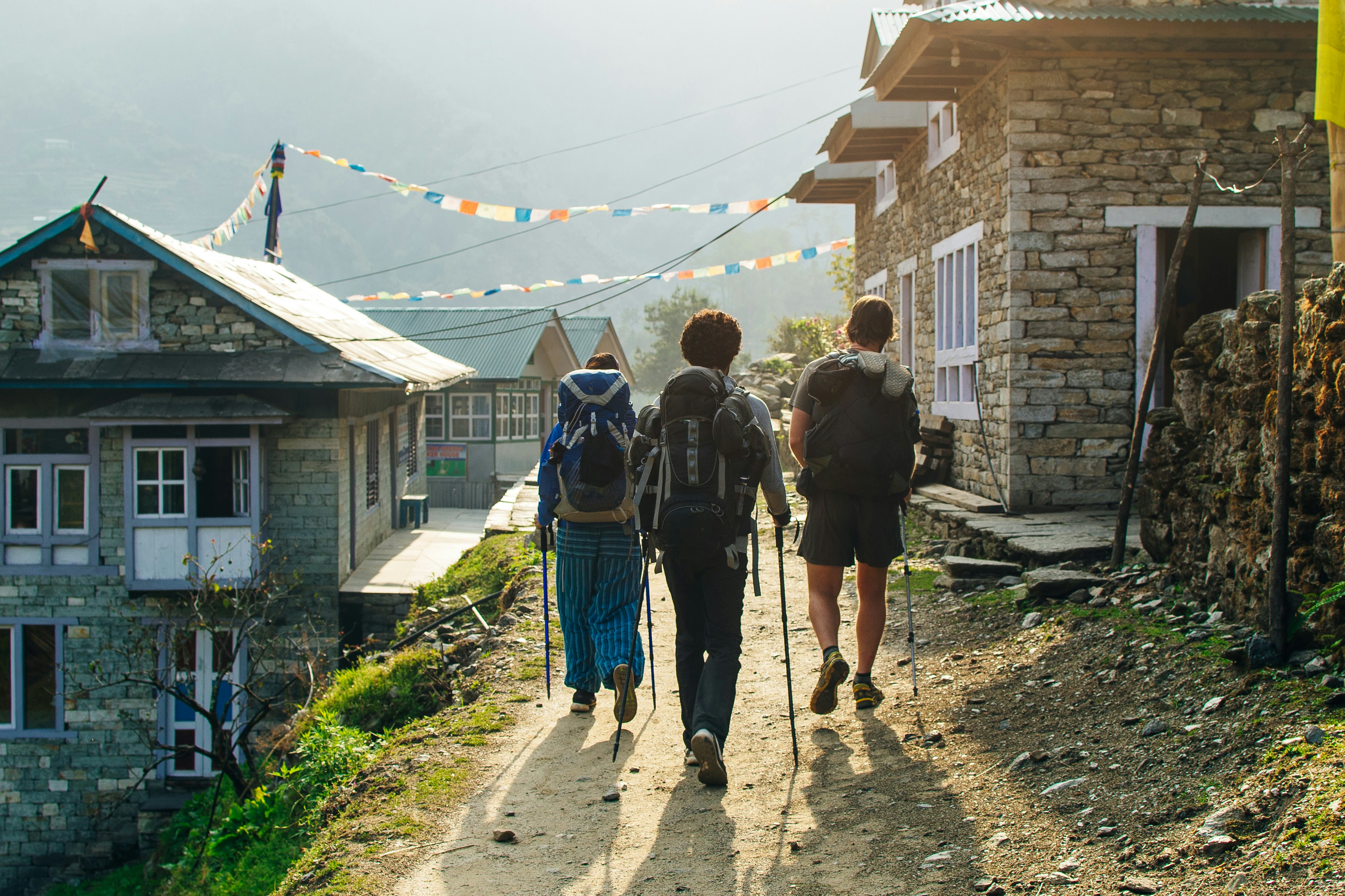 Three trekkers with backpacks and poles on a trail between brick houses on a steep hill. Colorful flags fly over head