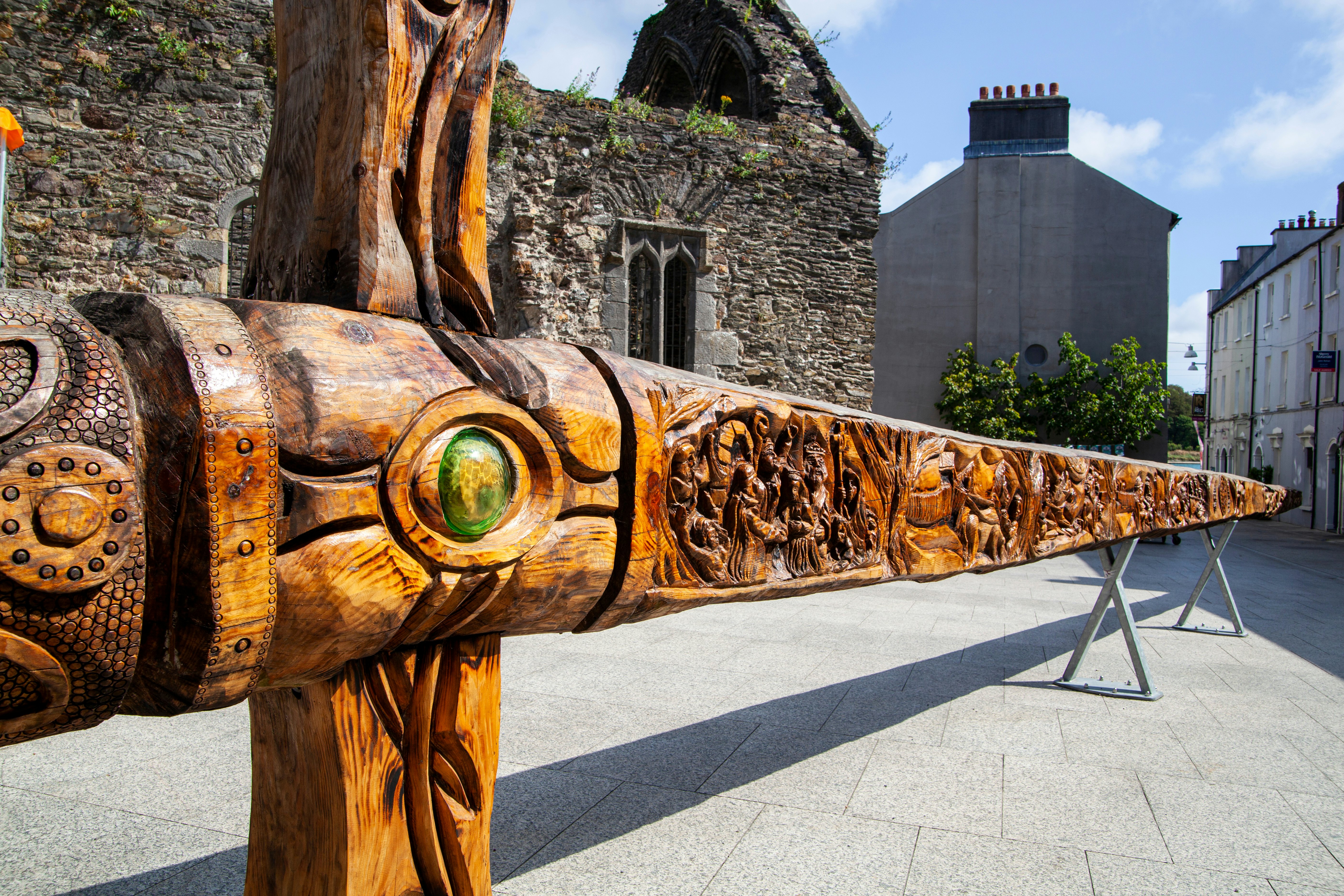 A view of a monumental wood sword with elaborate carvings on display in a city square. The ruins of a medieval building are seen behind the sword.