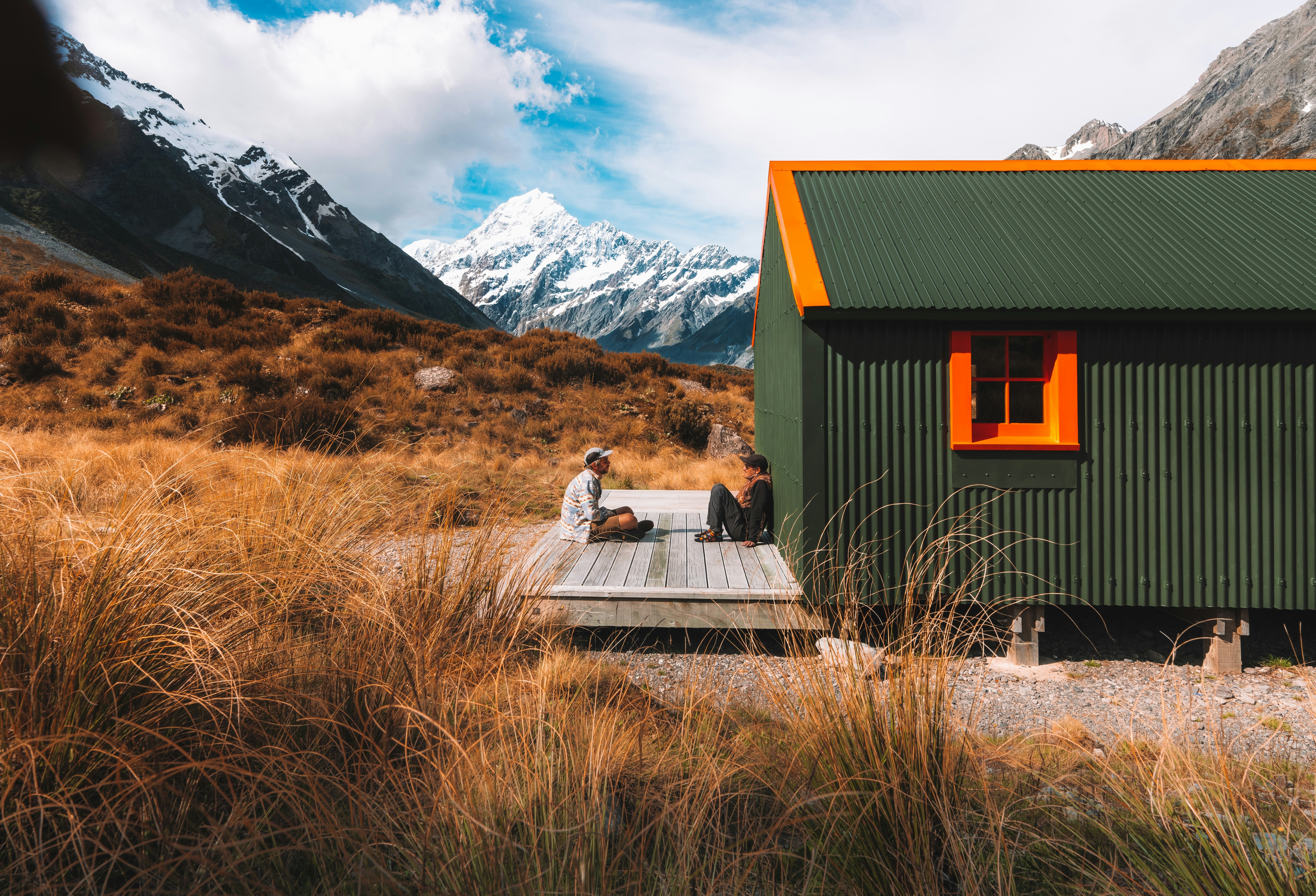 Two young male tourist traveler sit in front of a hut by mountains
