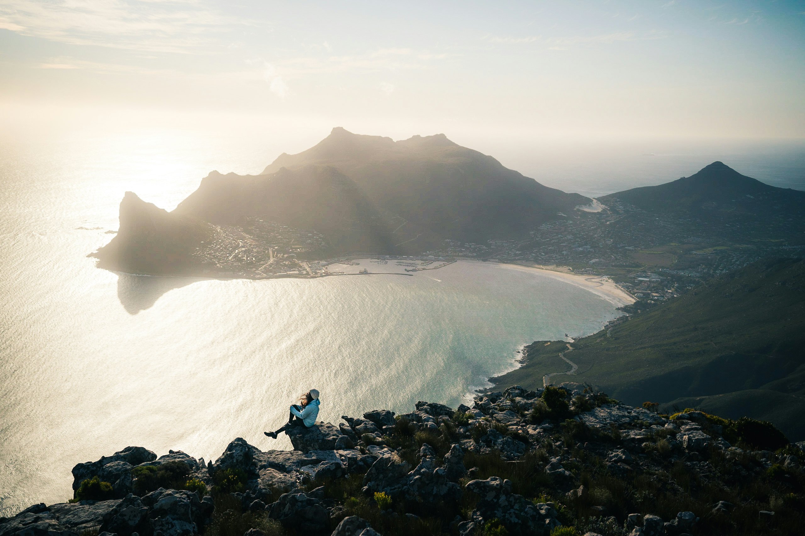 A silvery tint over a ocean with a woman seated on a rocky outcropping high above a cove with a beach and a mountain in the background.