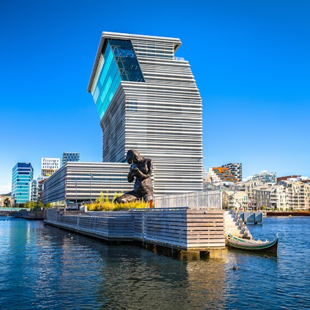Tall contemporary building, slanted at the top, surrounded by a harbor with a large human-formed sculpture in the foreground and city buildings in the far distance on a sunny day.