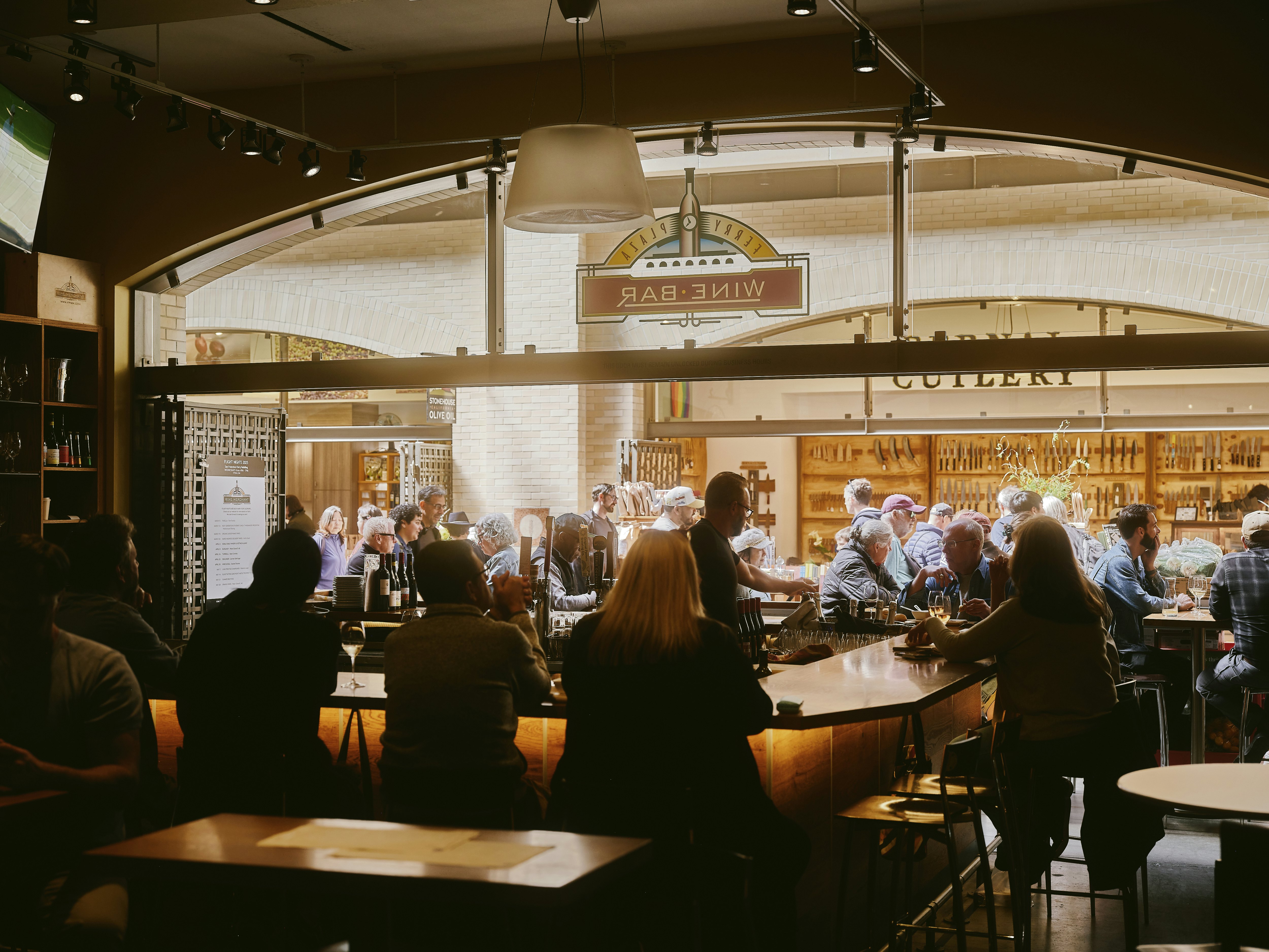 People bathed in golden light sitting around a u-shaped bar, enjoying drinks. More people waking past in the background
