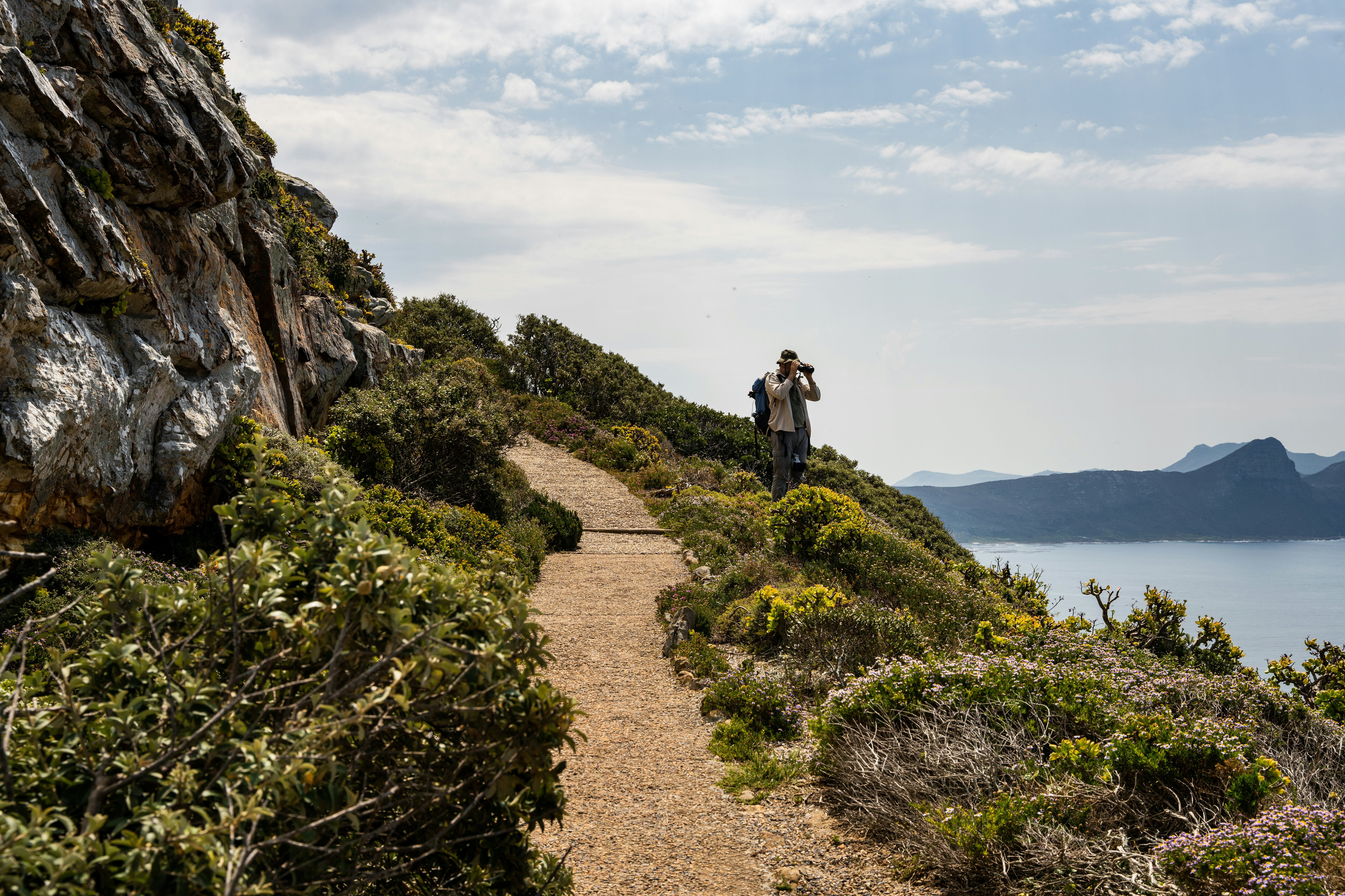 A man looks through binoculars on a narrow gravel path lined on either side with low brush. To the left is a rocky cliff to the right is a down slope to the ocean