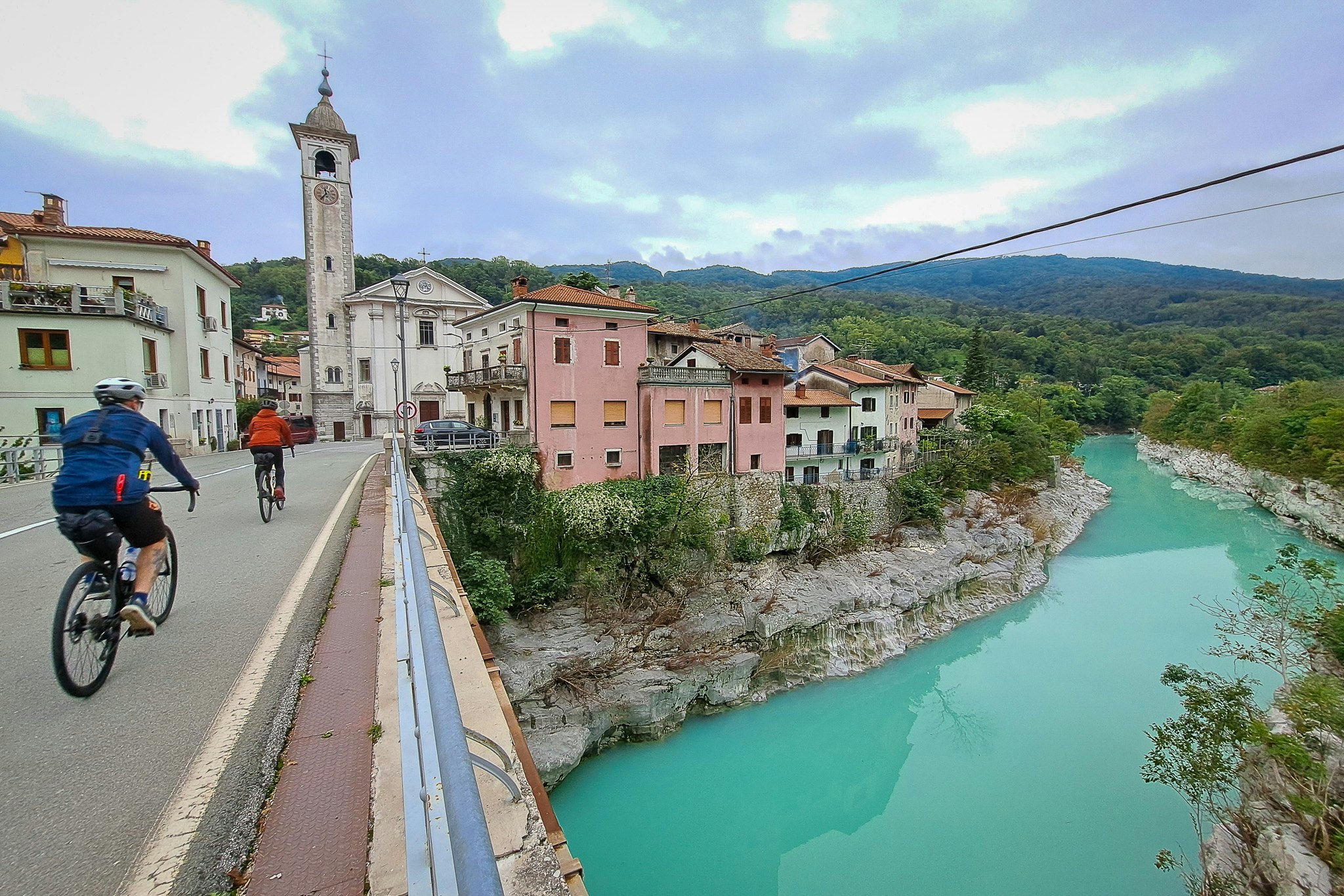 Two cyclists pedal bikes through a small town and over a bridge that crosses a turquoise river.